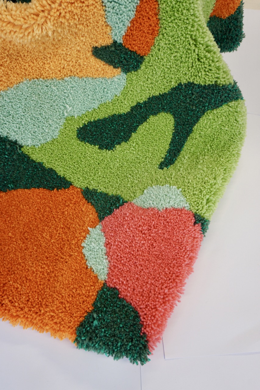 A textured rug featuring an abstract design with vibrant colours, including orange, green, light blue, and pink. The rug's fibres create depth and visual interest, with a bold combination of shapes and tones.