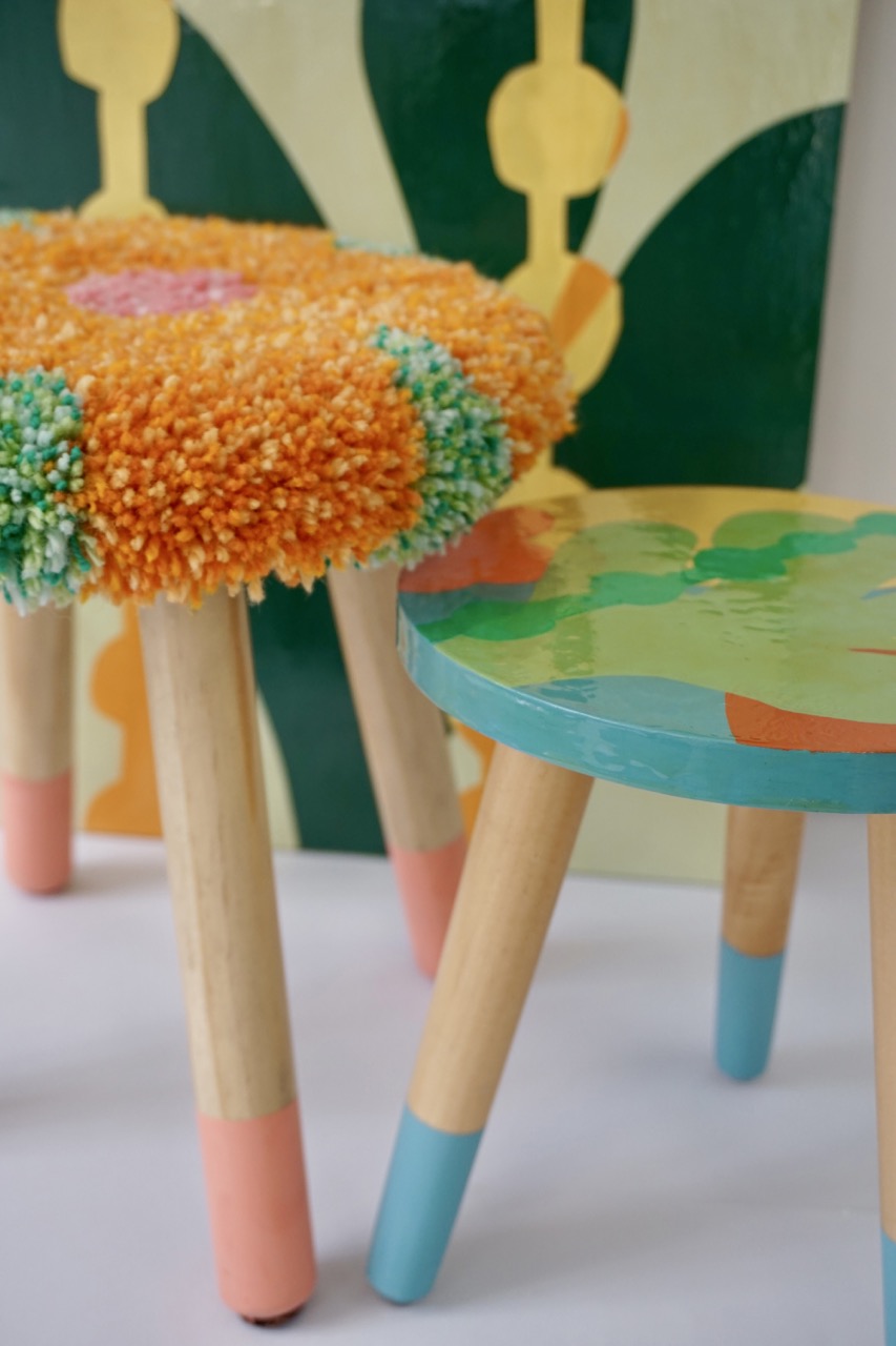 Two small stools are displayed on a white surface. The left stool has a fluffy floral top in orange and green, while the right stool features a painted surface in vibrant colours. Both stools have wooden legs with pastel-painted feet.