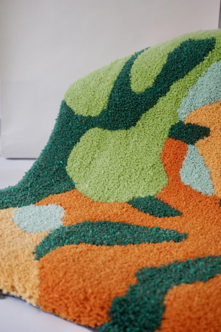 A textured rug featuring an abstract design with swirling shapes in shades of green, orange, and light blue. The rug is piled up slightly, showing its soft, fluffy texture on a neutral background.
