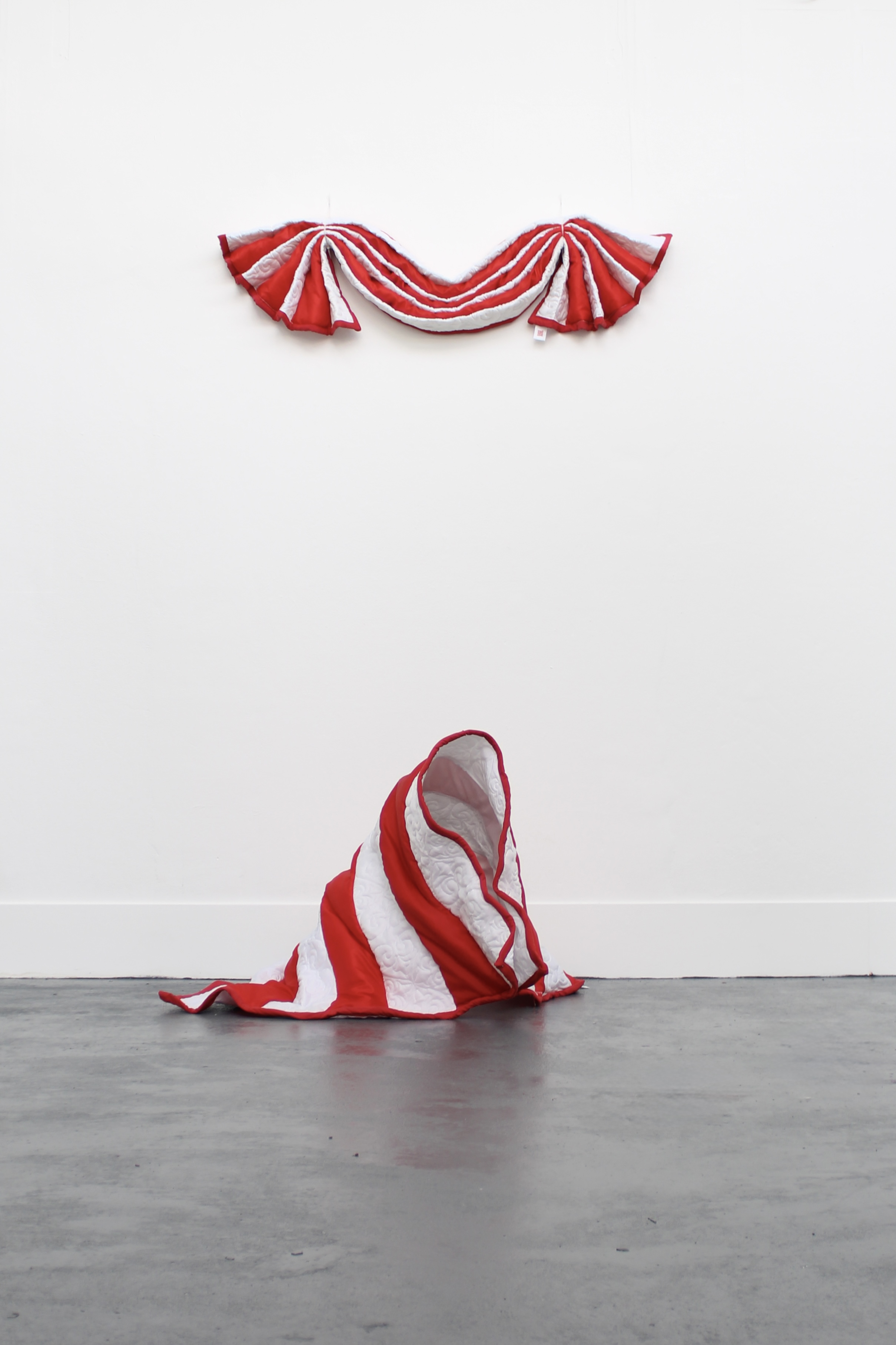 A large, red-and-white striped textile sculpture is hung on the wall above a crumpled version of the same pattern lying on the floor. The background is a plain white wall, and the flooring appears to be a grey concrete.