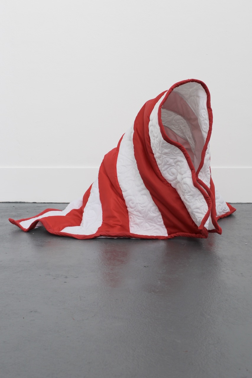 A red and white striped quilt draped haphazardly on the floor, with the fabric forming an abstract shape that suggests a hood or a cloak. The background is plain and the floor is dark, providing contrast to the quilt.