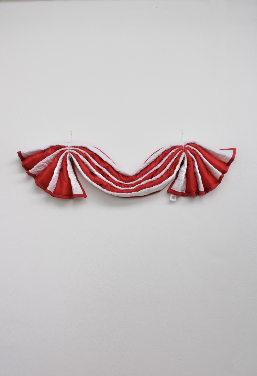 A decorative fabric drape featuring alternating red and white pleats, arranged in a wave-like manner. The fabric is suspended against a plain background, showcasing its pleated design.