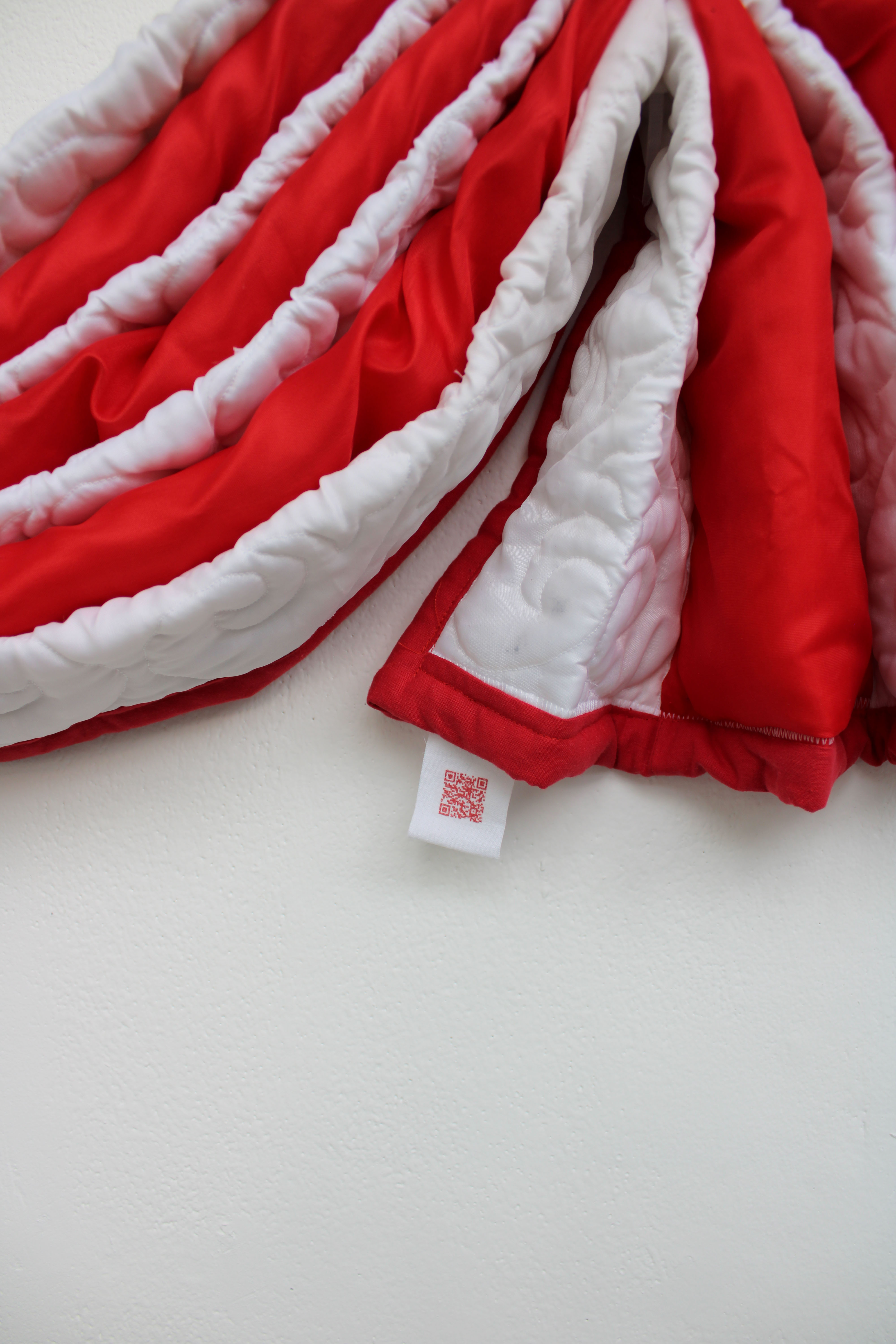 A red and cream quilt draped over a white wall, showcasing its textured surface. The edges are neatly finished, with a small label visible at the bottom right corner.