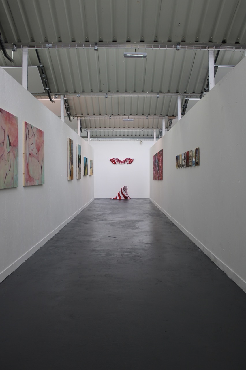 A long, narrow art gallery with white walls, showcasing colourful paintings on both sides. At the far end, a sculpture draped in red and white stripes is positioned on the floor, beneath a metal roof with exposed beams.