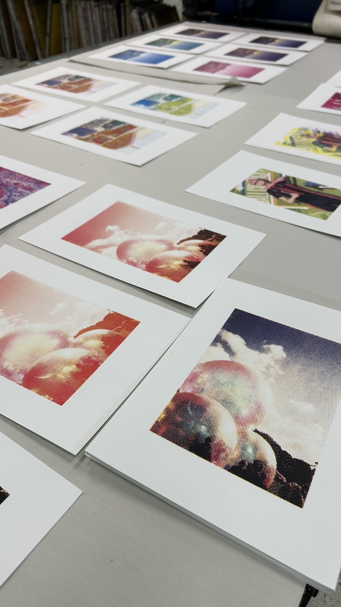 Printed artworks spread on white table surface. Foreground shows red, orange, and blue abstract compositions. Background displays various coloured prints in grid arrangements.