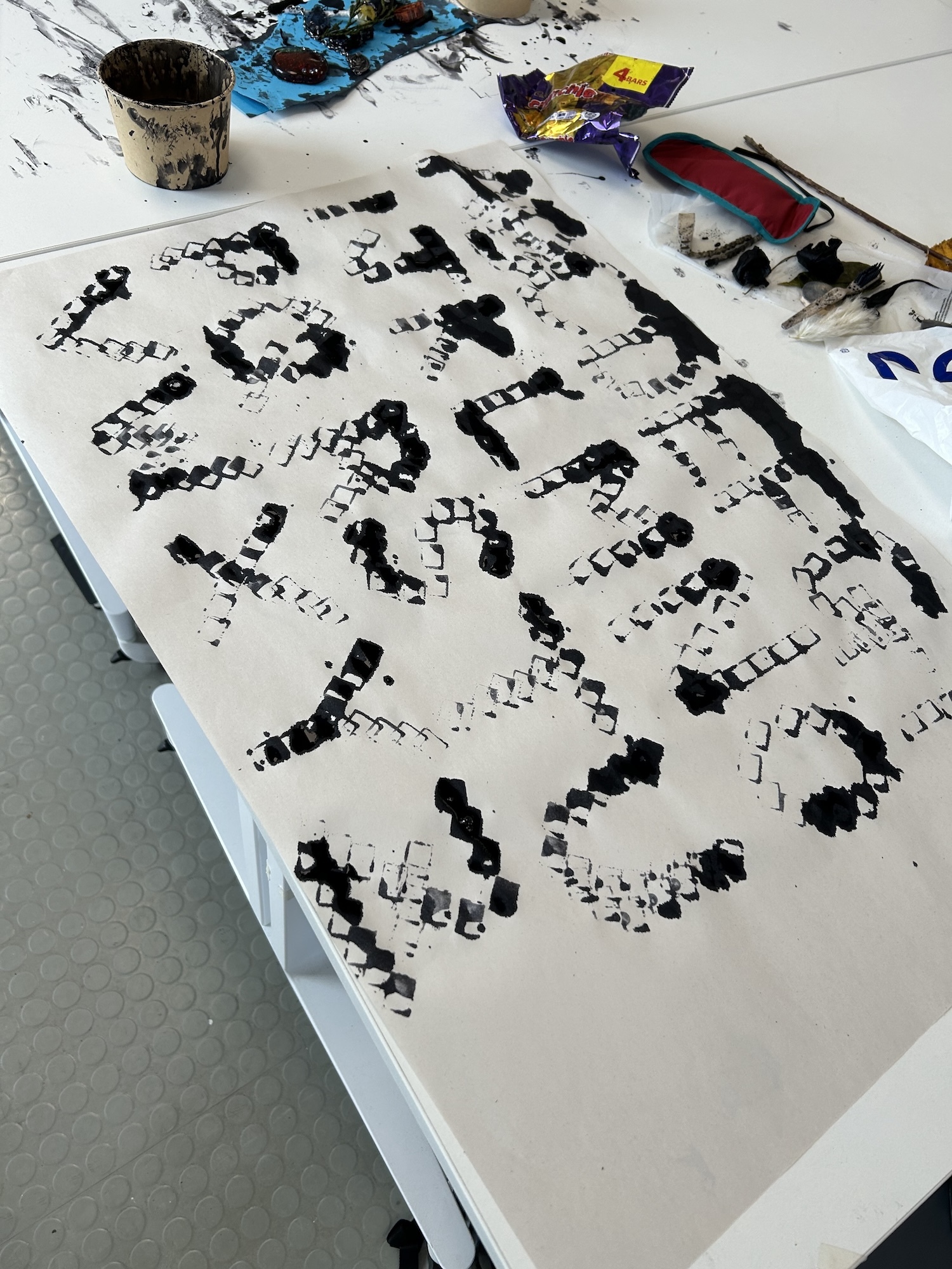 Black geometric pattern on white paper with pixelated skull design, paint pot and art supplies visible on table surface.