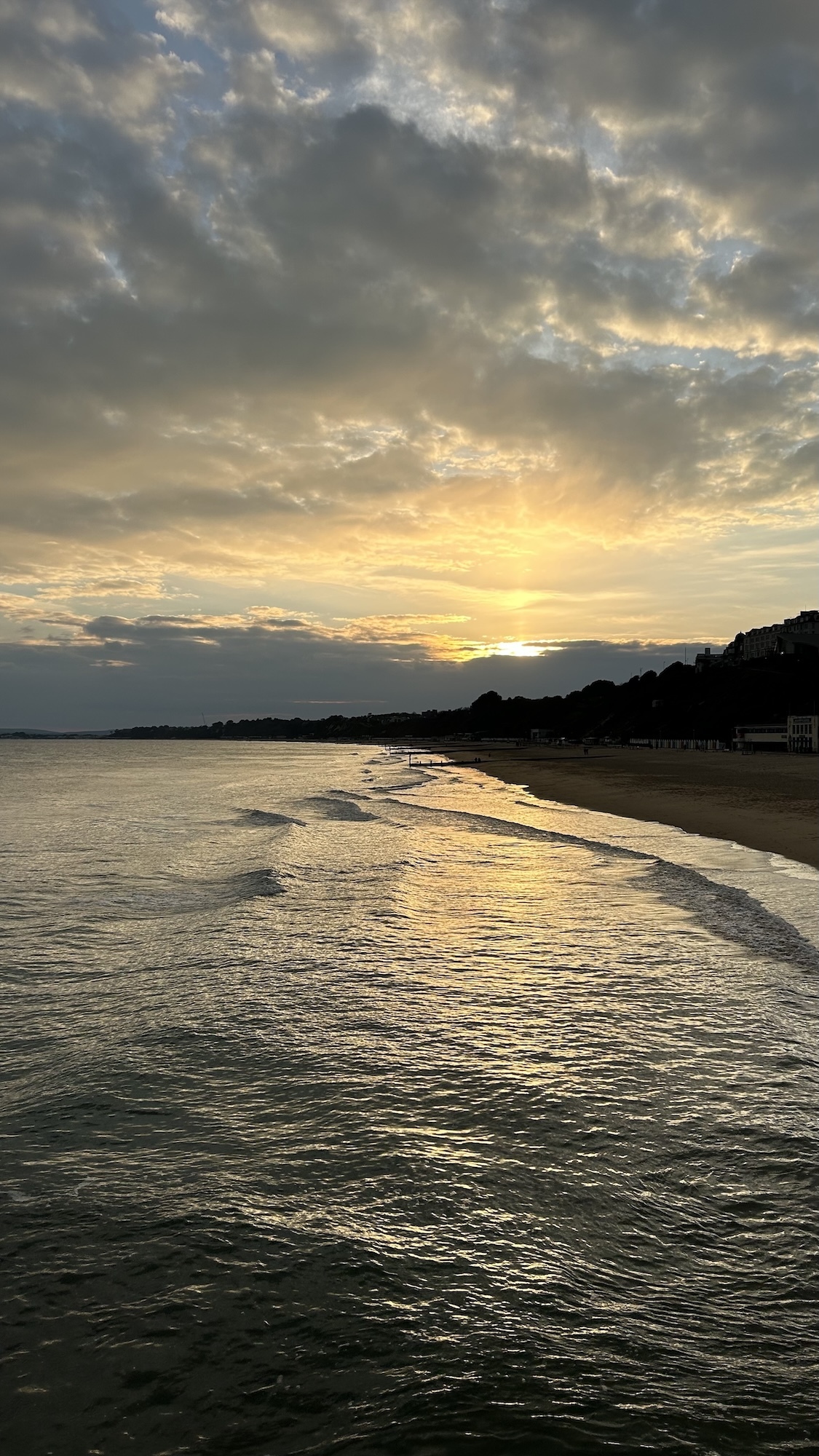 Dramatic cloudy sky with golden sunset light over ocean waves meeting sandy beach, dark silhouetted coastline on right side.