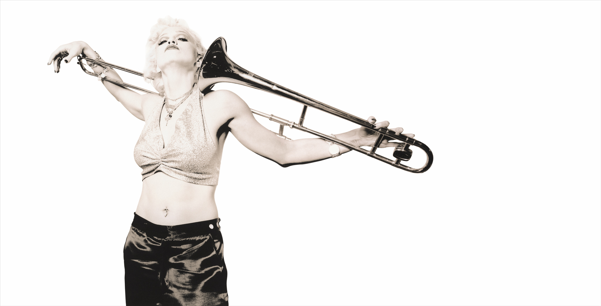 Madonna holding trombone horizontally above head, wearing white top and dark leather trousers against white background.