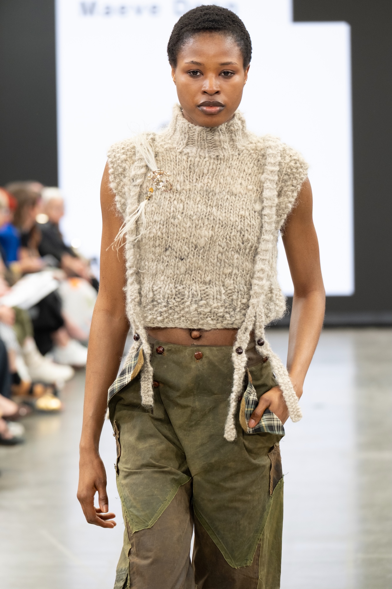 A model walks down the runway wearing a sleeveless, knitted beige top with a high collar and loose threads. She pairs it with green cargo trousers, featuring buttons and patchwork details, while the audience is visible in the background.