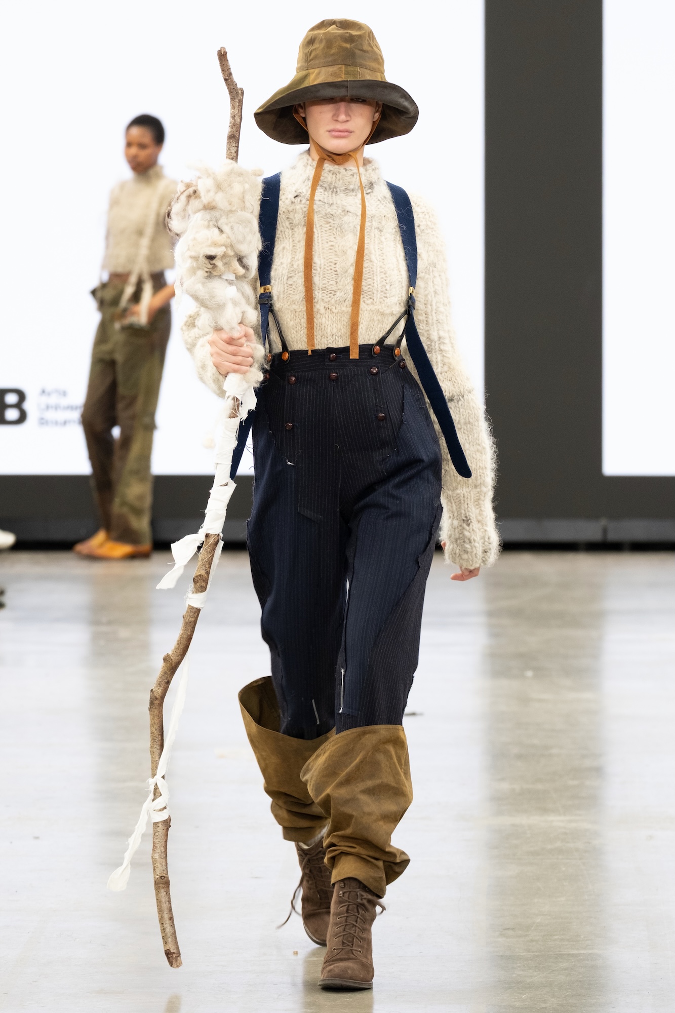 A model walks down the runway at Graduate Fashion Week wearing a beige knitted sweater, dark overalls, and brown ankle boots, holding a stick. They are accessorised with a wide-brimmed hat and the background features large screens displaying text or images.