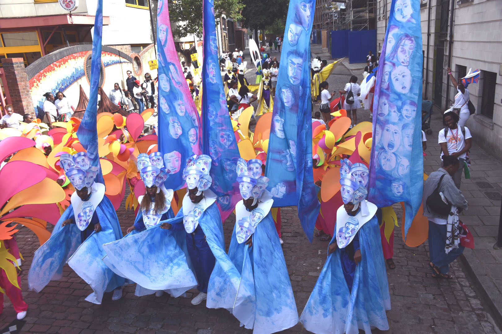 Four carnival performers lined up wearing blue and white costumes, with headdresses and banners behind them. The headdresses and banners have faces illustrated onto them.