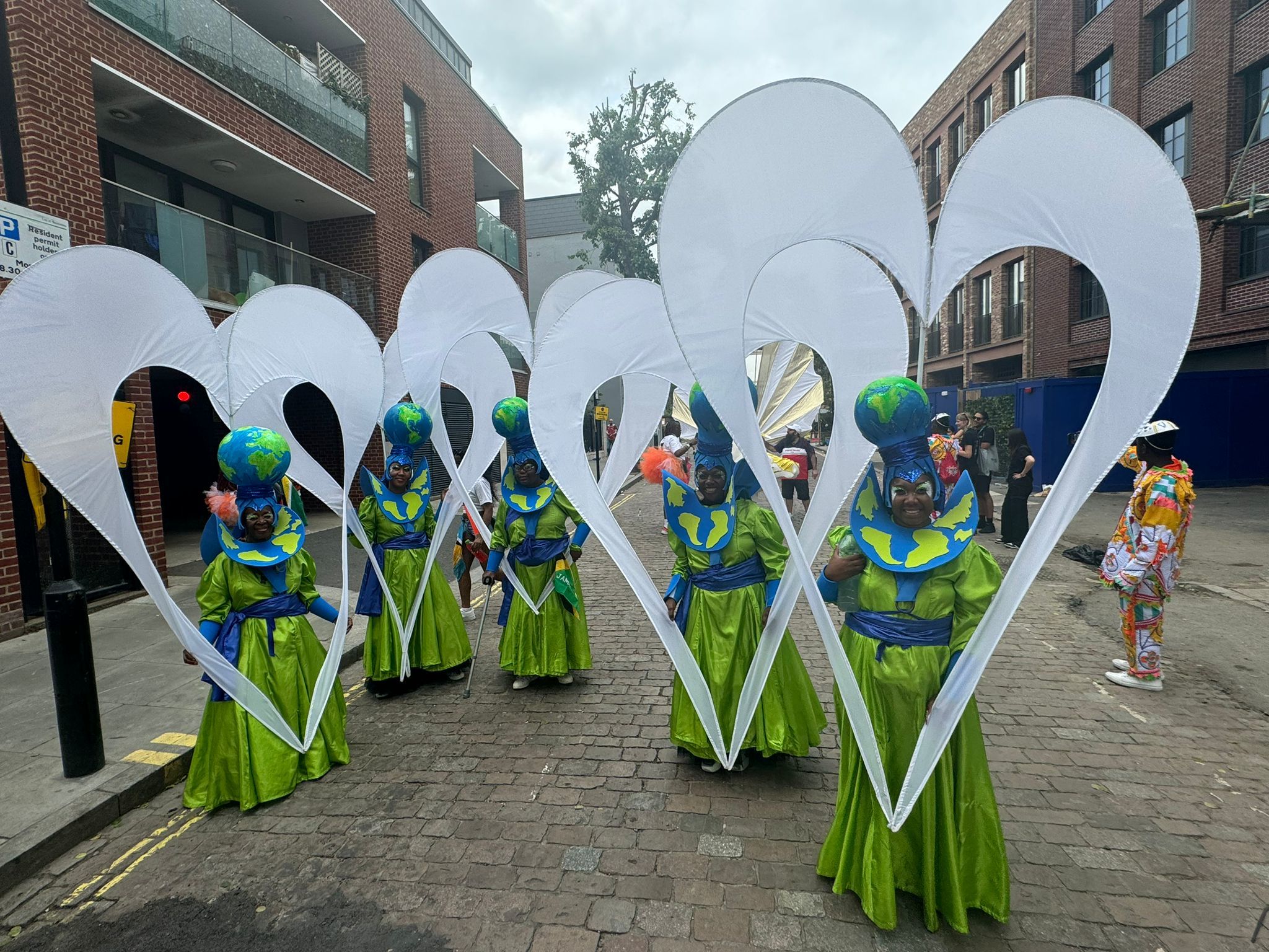 Carnival performers in green and blue Earth-themed costumes holding giant white hearts performing on a street.