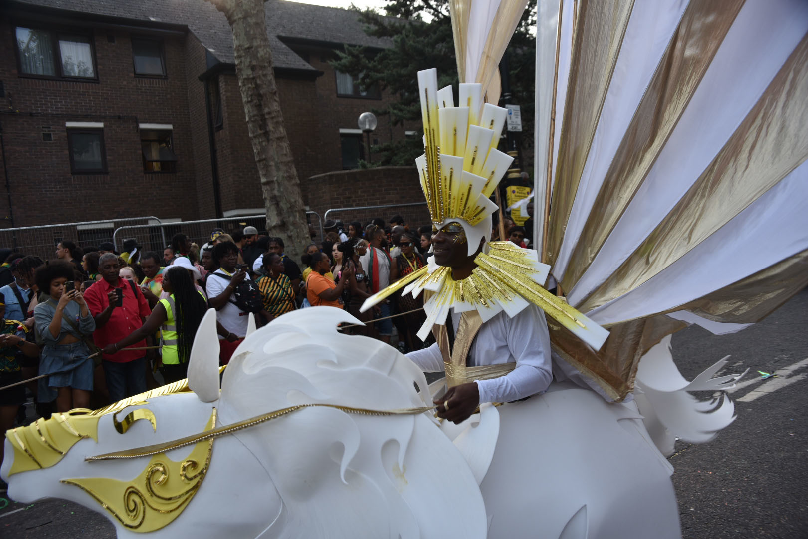 A carnival performer wearing a white and gold costume and headdress, with a white and gold horse puppet in front of them.