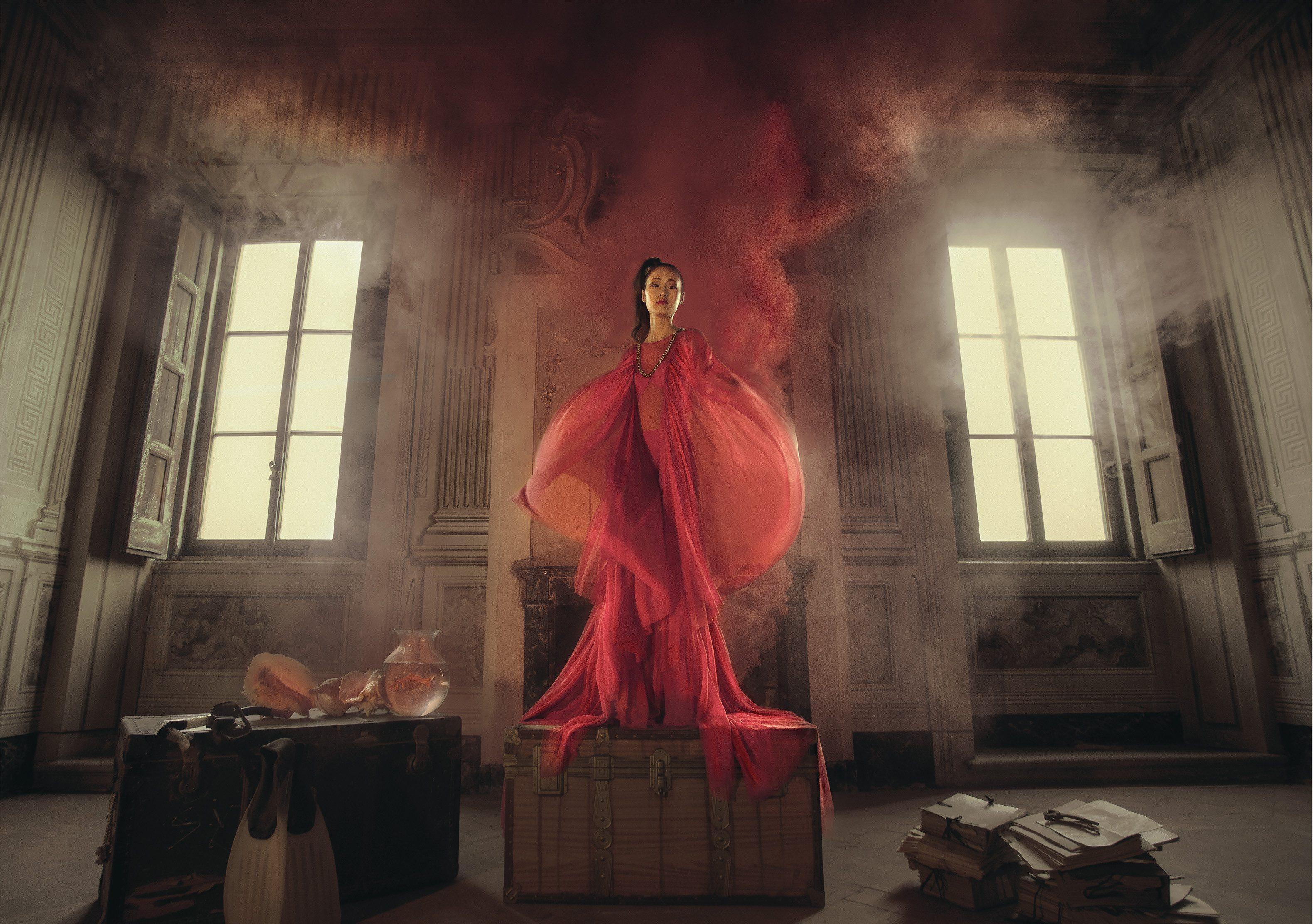 Lady in red dress stands on a chest with smoke coming out from behind her. There is a luggage trunk to her right and a pile of documents to her left.