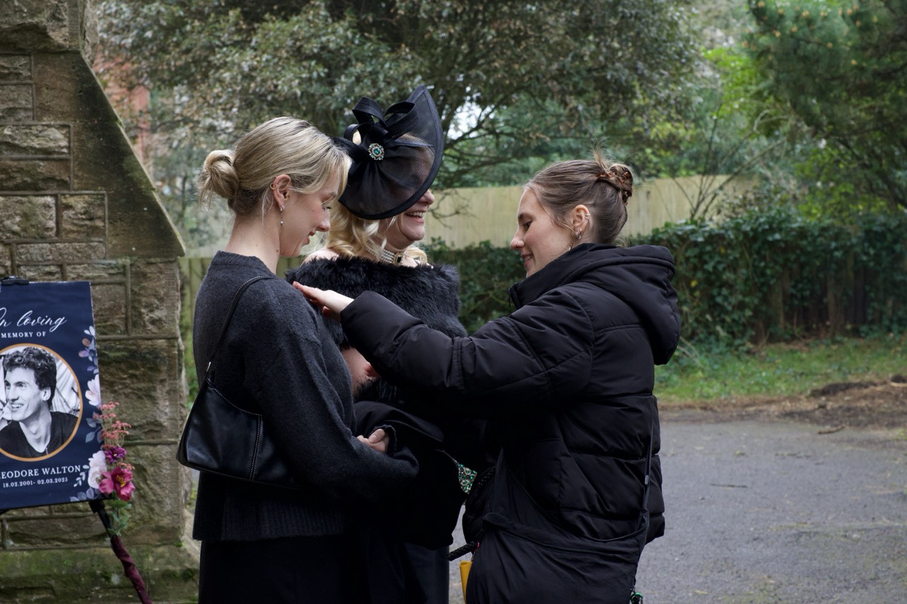Three women are gathered outdoors near a stone wall. One woman in a black dress adjusts the clothing of another woman wearing a black hat and coat. The background features greenery and a poster with a photo of a man.