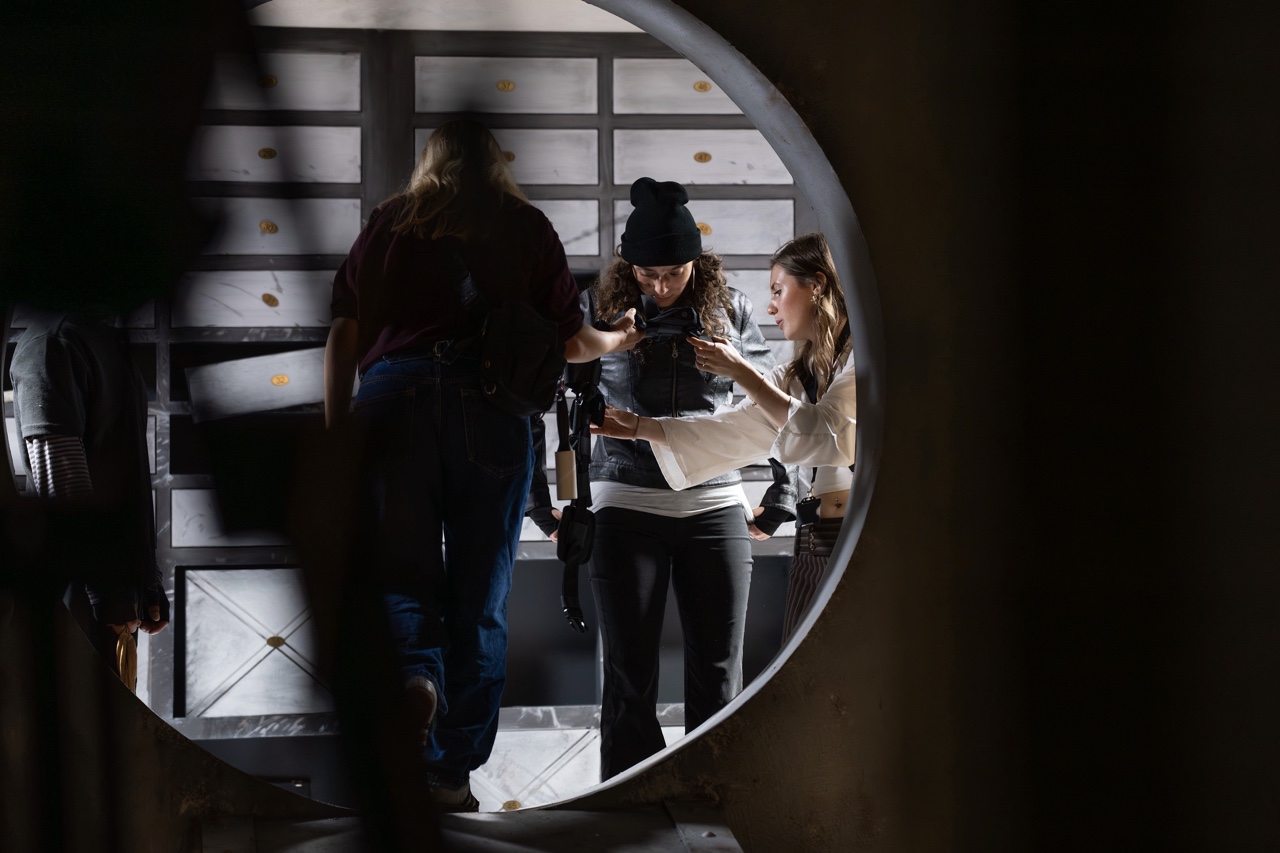 A group of three people is seen from a circular opening in a structure. One woman, wearing a black beanie and a white long-sleeved top, is engaging with another woman, while a person in blue jeans stands behind them. The background features grey panels with spots.