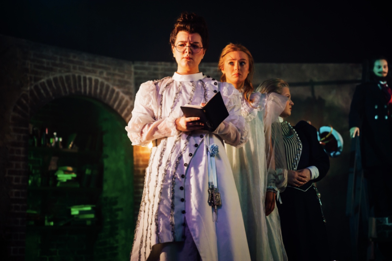 A group of actors on stage during a theatrical performance. The central figure, a woman in a white costume holding a book, stands in front, flanked by two other women in pale dresses, with a darkened scene and shelves in the background.