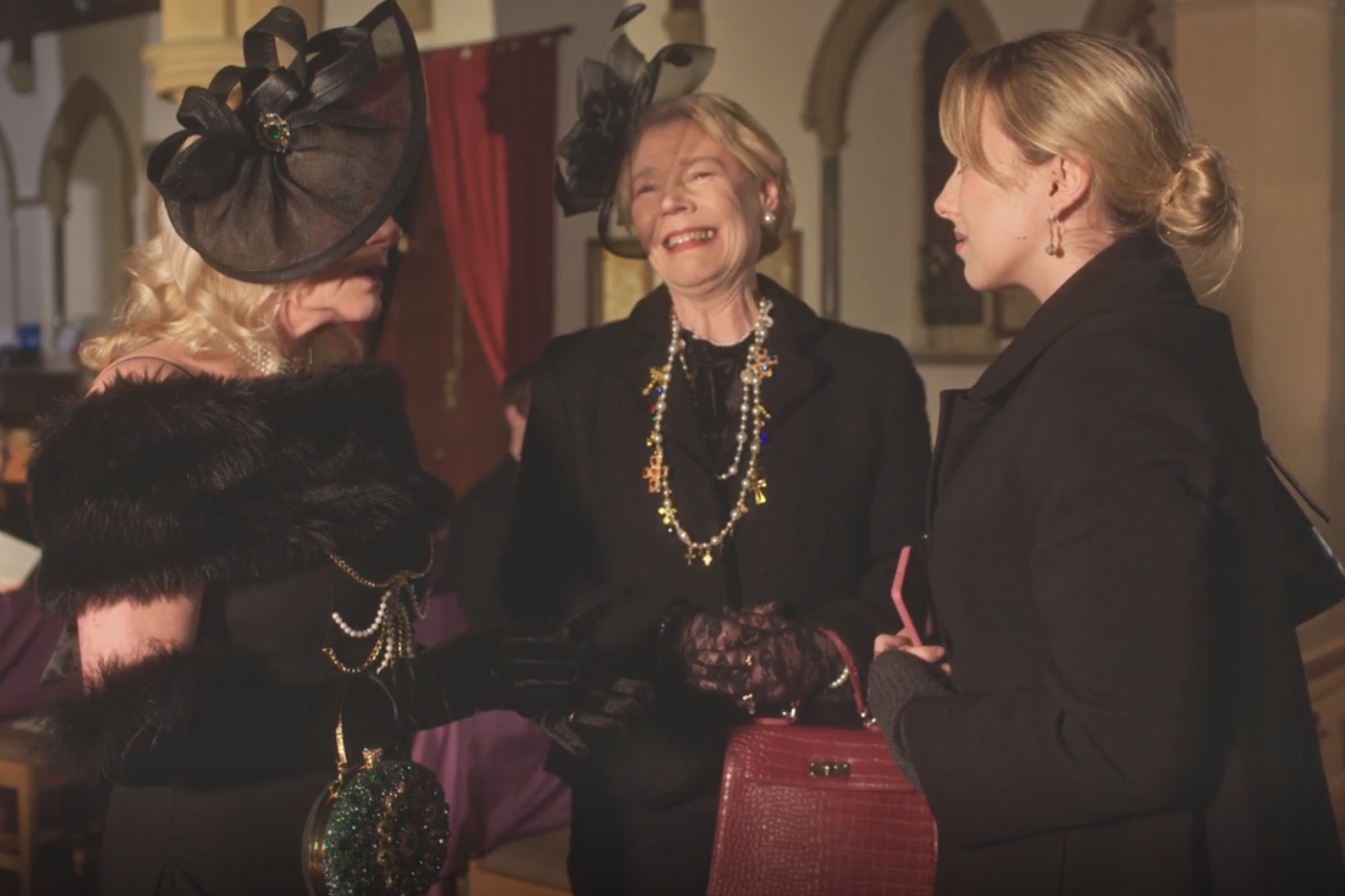 Three women in elegant attire engage in conversation. One woman wears a large black hat and fur stole, adorned with a beaded handbag. The second woman has short hair and wears a pearl necklace, while the third woman holds a pink handbag and has long hair, dressed in a coat.