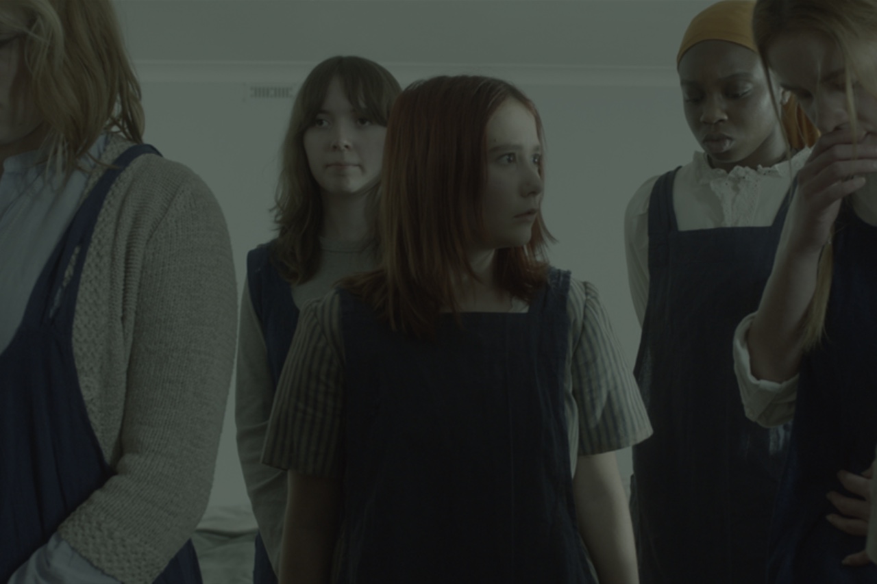 A group of five women in dark pinafore dresses stands closely together in an indoor setting. Some women have long hair and varying skin tones, while one woman wears an orange headscarf. They are positioned against a light-coloured wall.