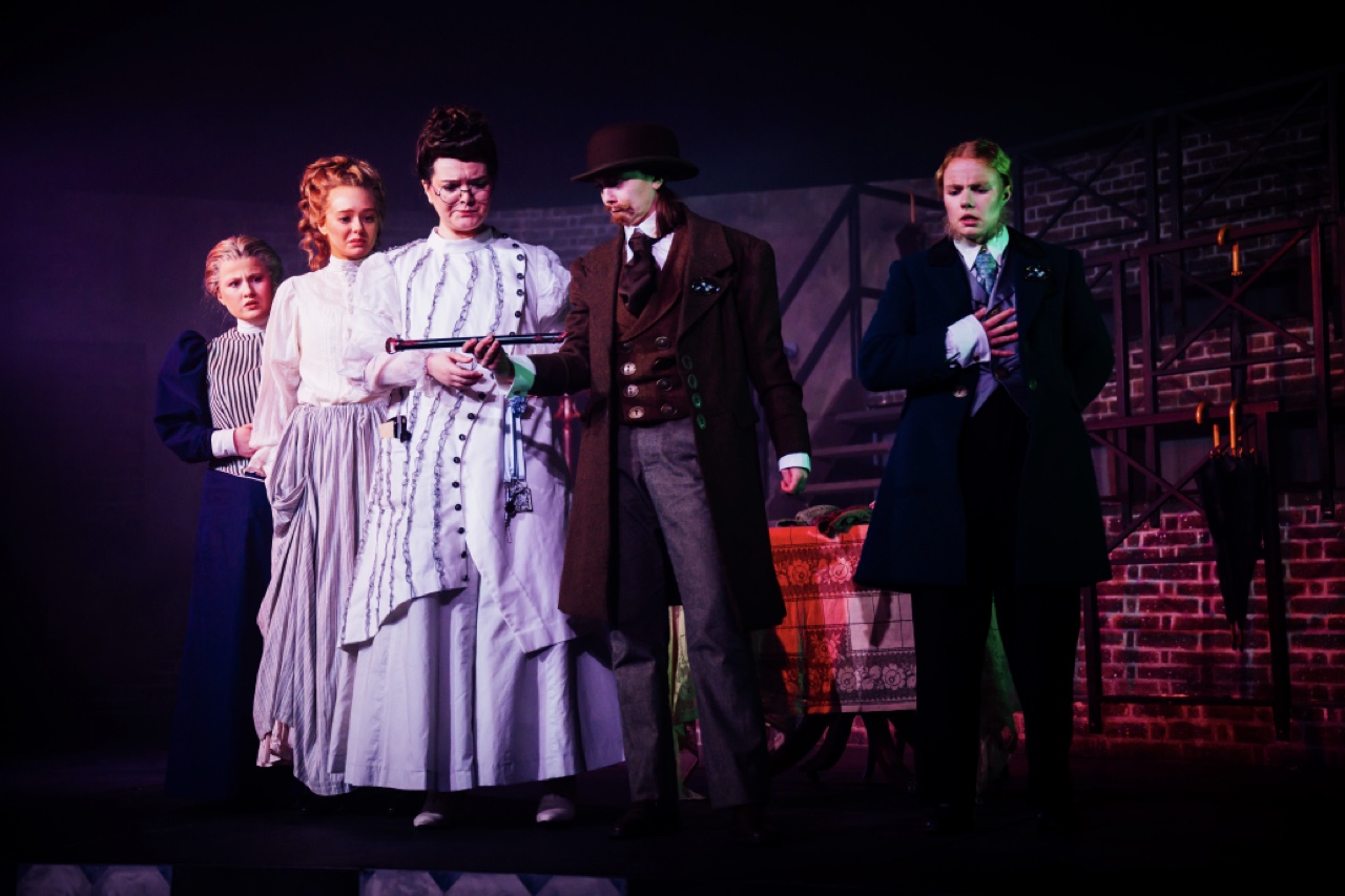 A group of five actors on stage in period costumes, featuring a woman in a white dress with a black shawl, a man in a brown coat and top hat, and three others in formal attire. The background includes a brick wall and dim lighting, creating a theatrical setting.