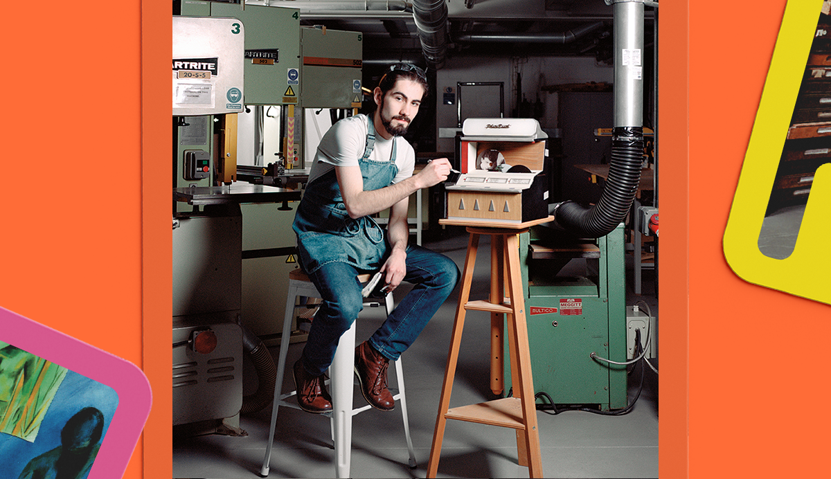 Bearded man in denim overalls sits on white stool in workshop with green machinery, orange walls, and colourful geometric borders.