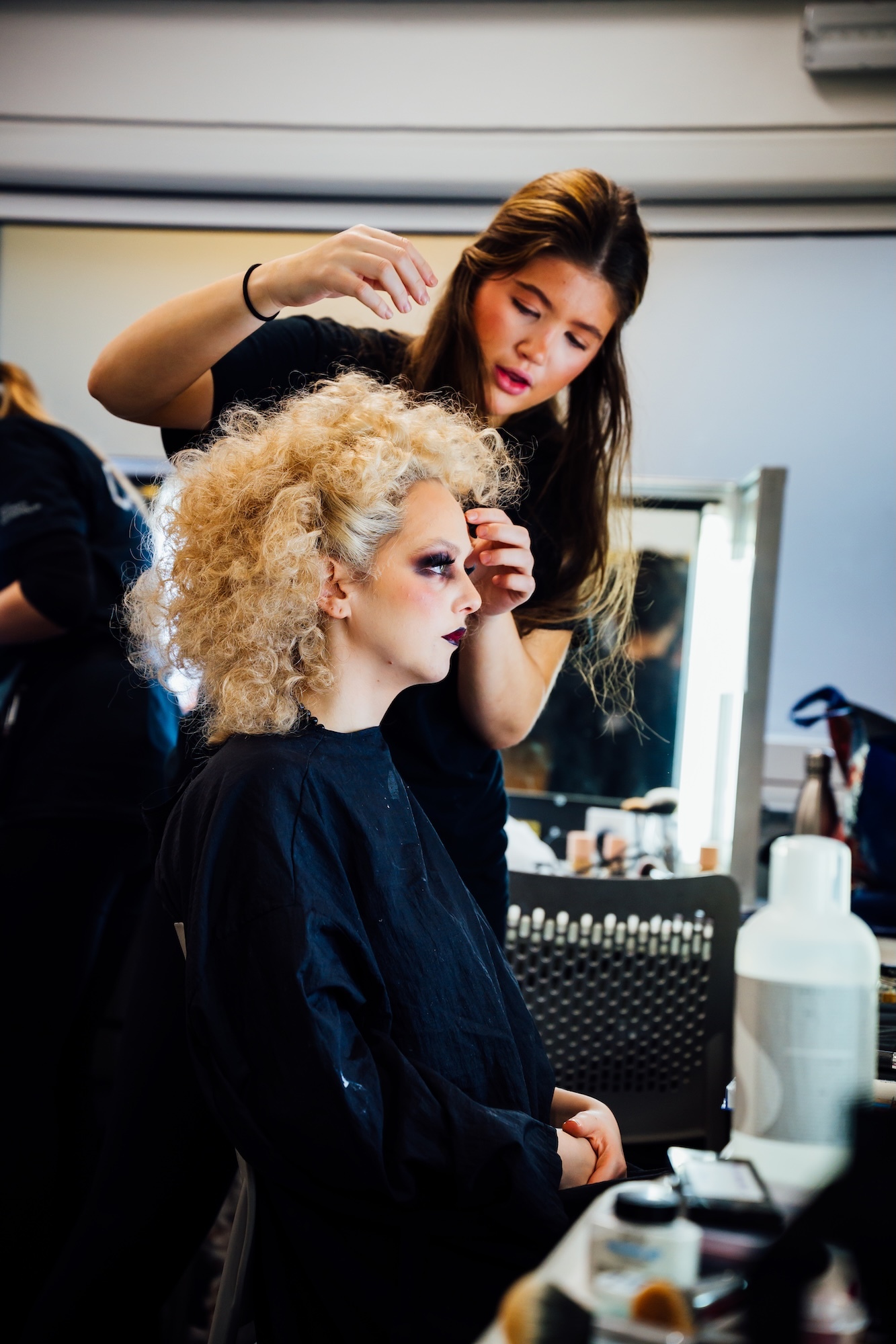 BA (Hons) Make-up, Hair and Prosthetics student applying cosmetics to a BA (Hons) Acting student with curly blonde hair in black cape, seated at mirror with beauty products visible.