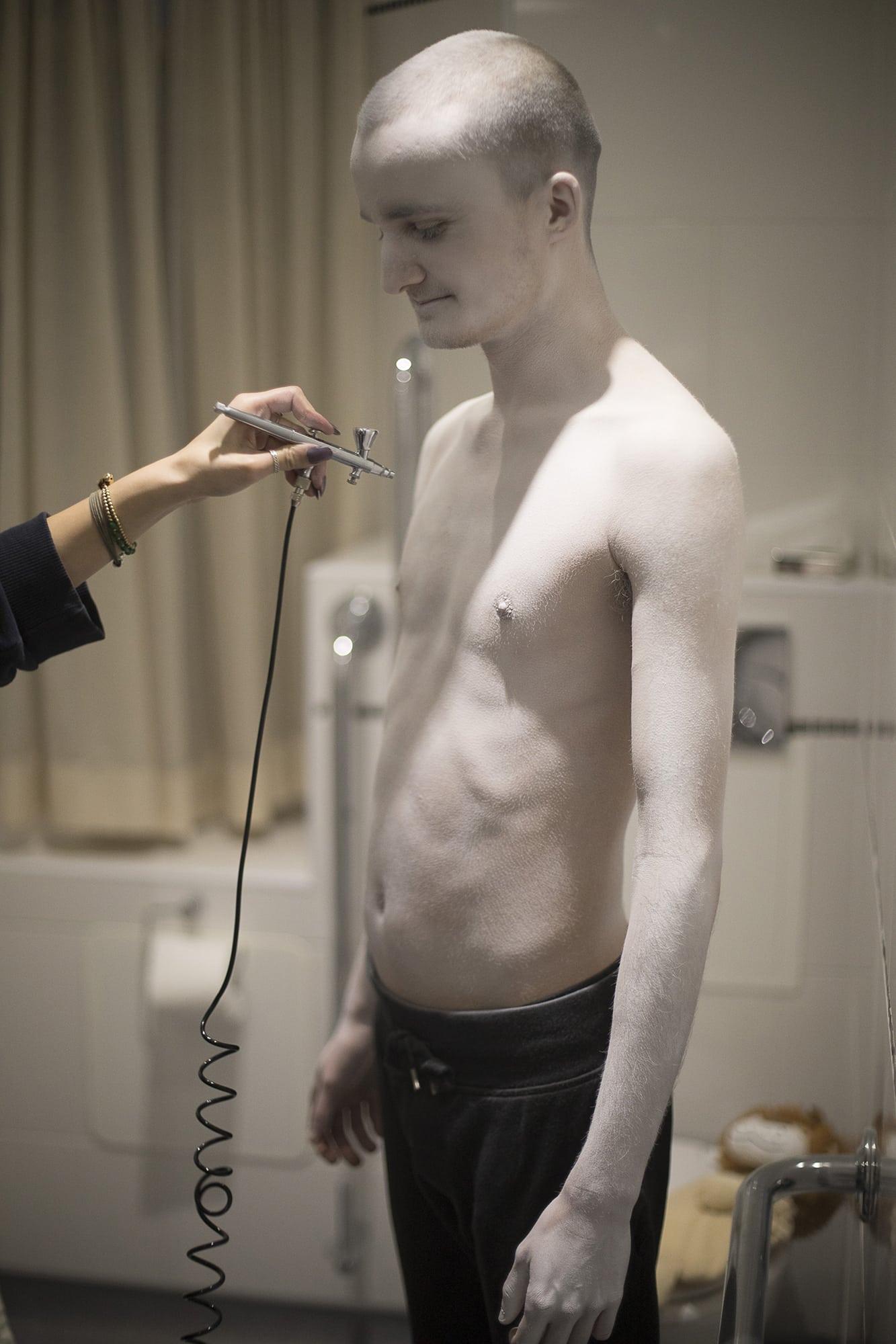 A man wearing no shirt stands while someone holds a metal instrument towards them.