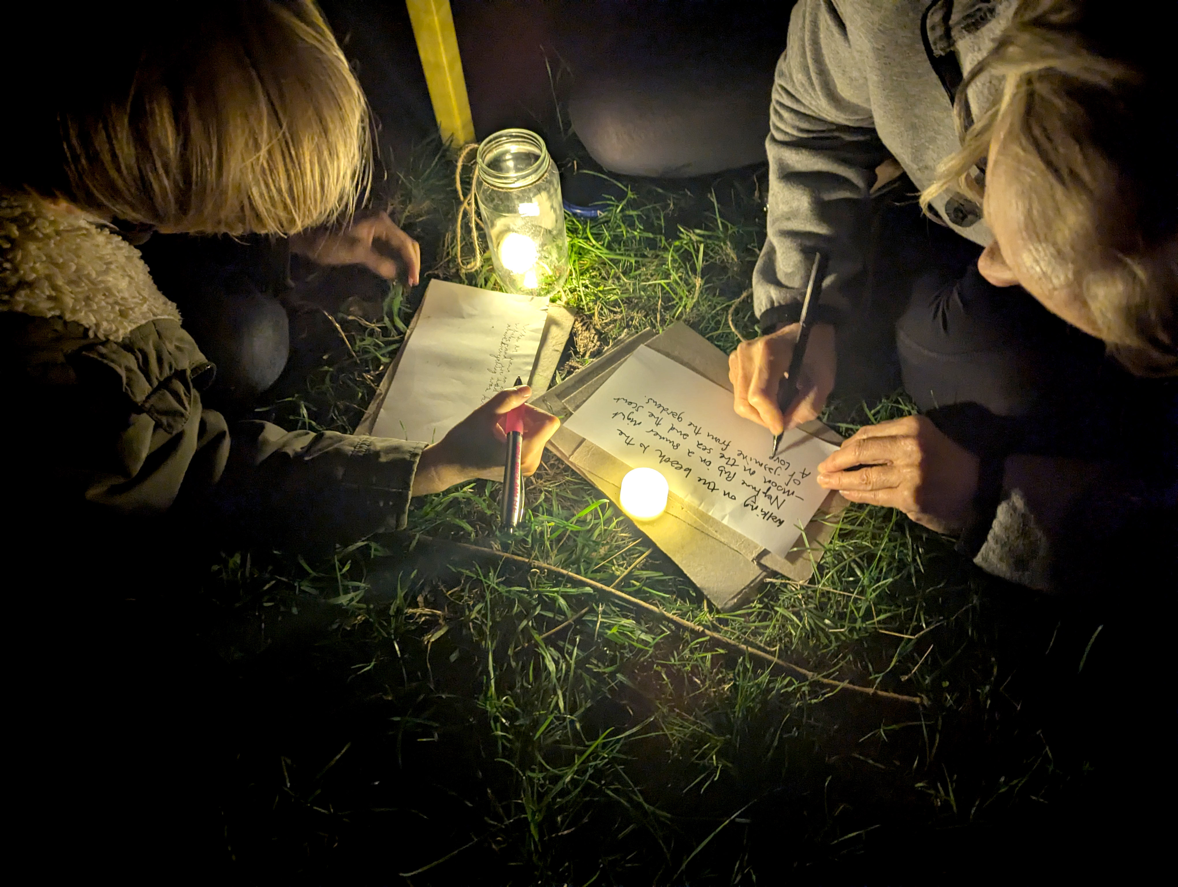Two people write on sheets of paper on grass at night, lit by a jar lantern and a small candle.