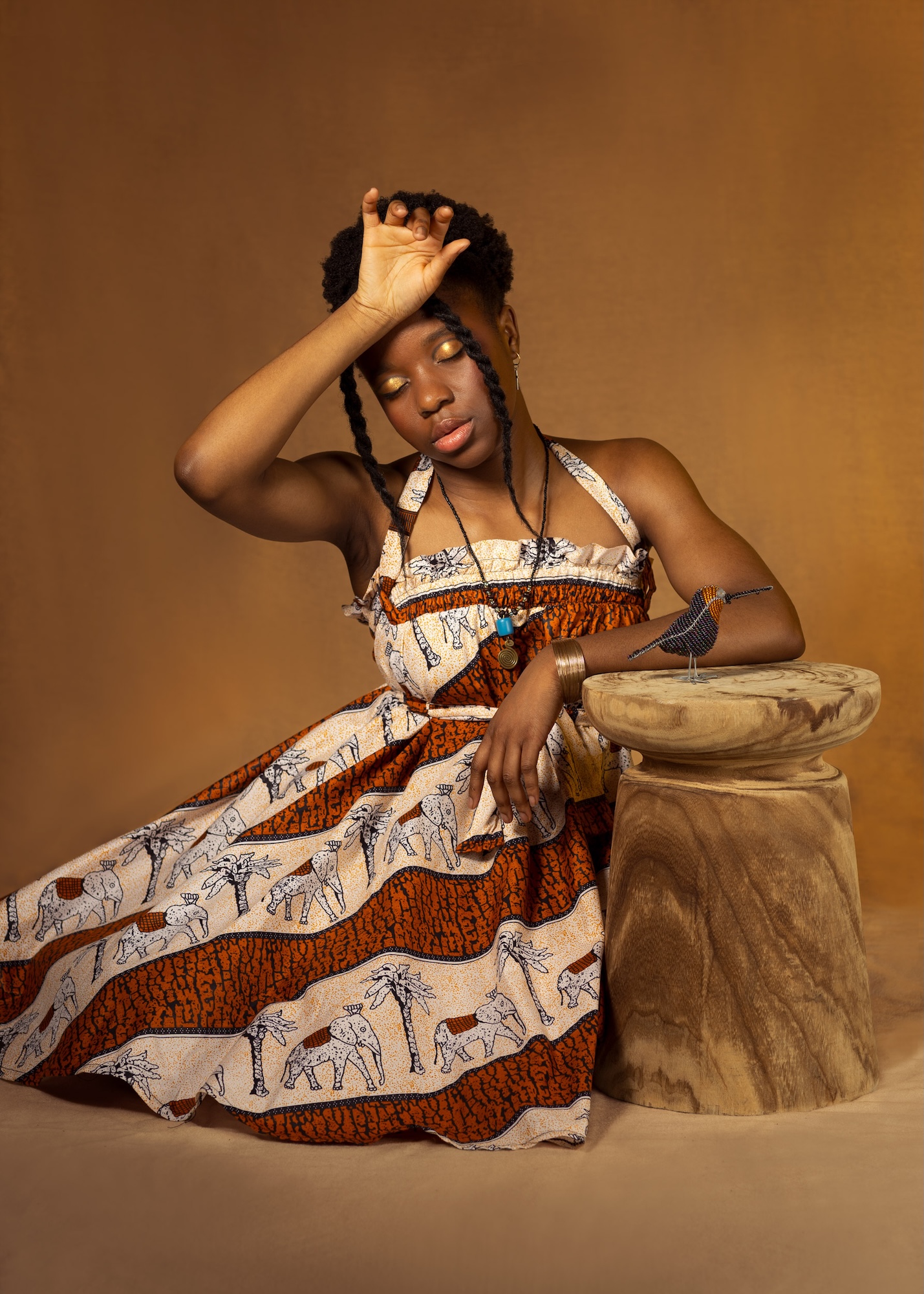 A fashion model wearing a white and brown dress with an elephant and palm tree pattern sits and poses, eyes closed, one hand raised to their forehead, next to a pedestal with a small model bird on it