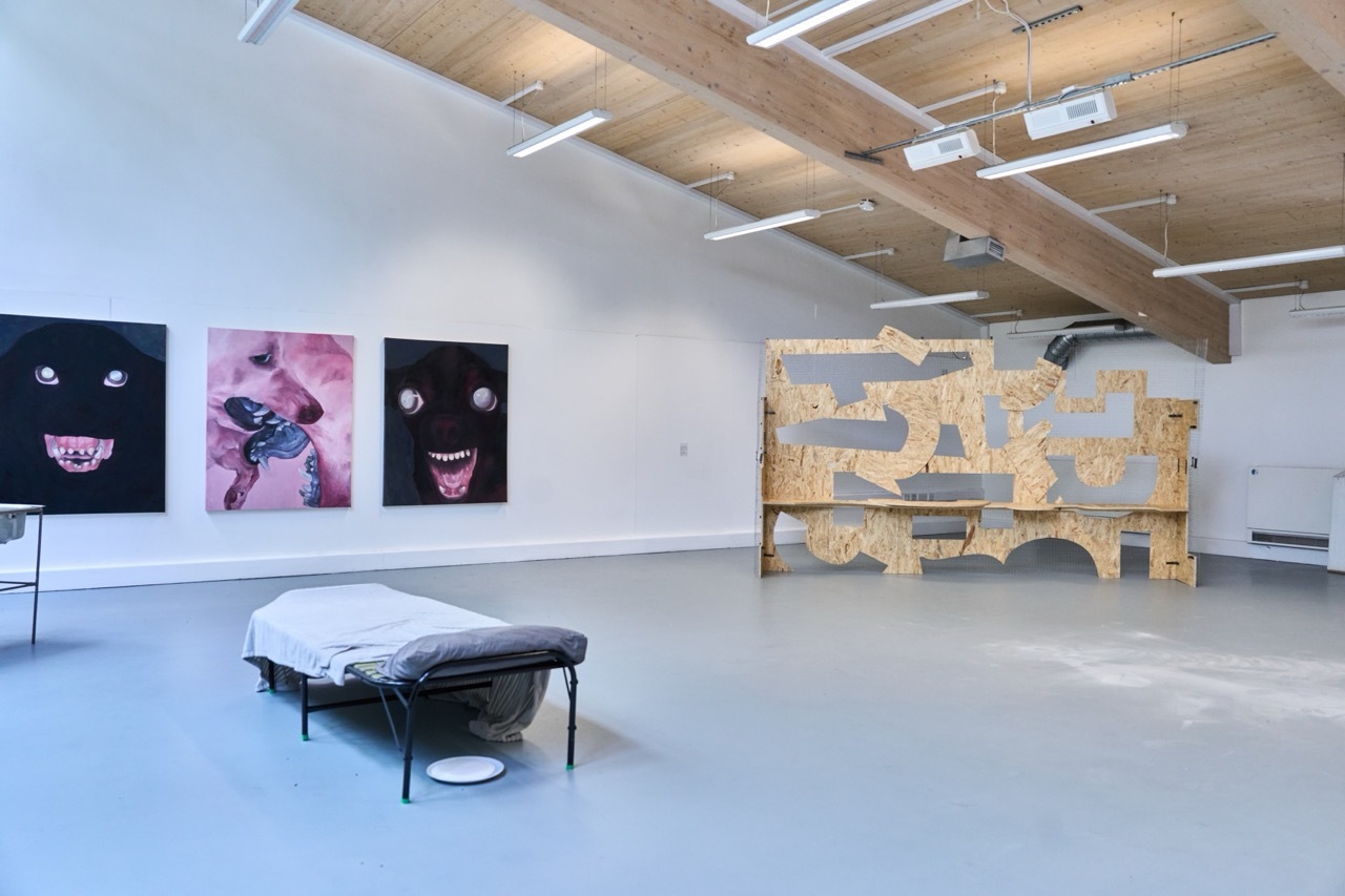 Gallery space with white walls, exposed wooden ceiling beams, three dark artworks with pink accents, wooden sculptural installation, and black bench on polished concrete floor.