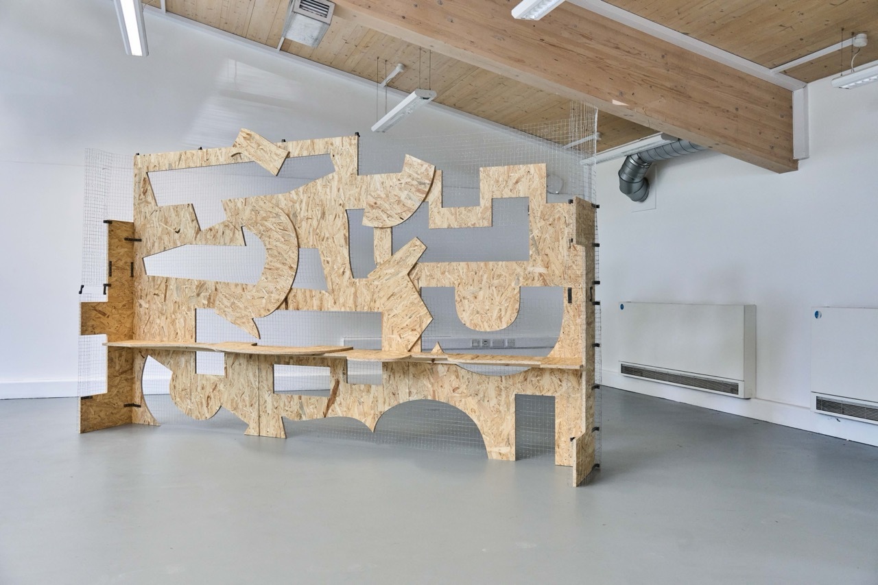 Large geometric wooden sculpture with interlocking angular forms in white gallery space with exposed ceiling beams and concrete floor.