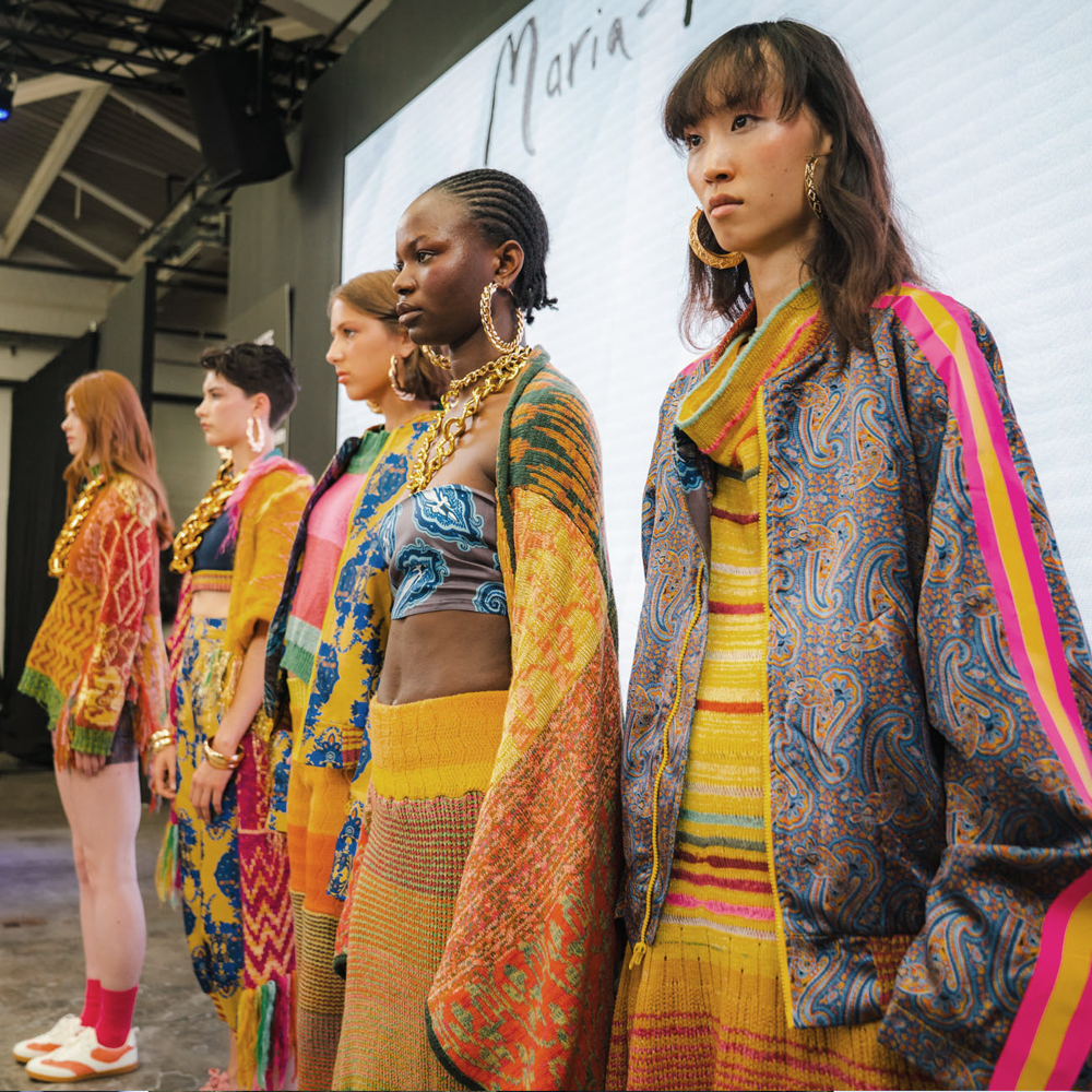 Models on catwalk wearing colourful patterned garments in yellow, orange, pink and blue with geometric and floral designs.