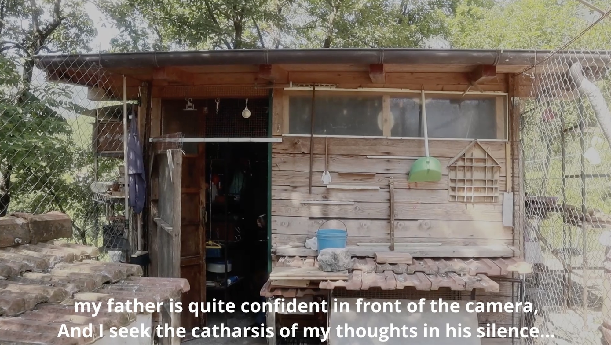 Film still of a small wooden shack or shed. Subtitles along the bottom read "my father is quite confident in front of the camera, and I seek the catharsis of my thoughts in his silence..."