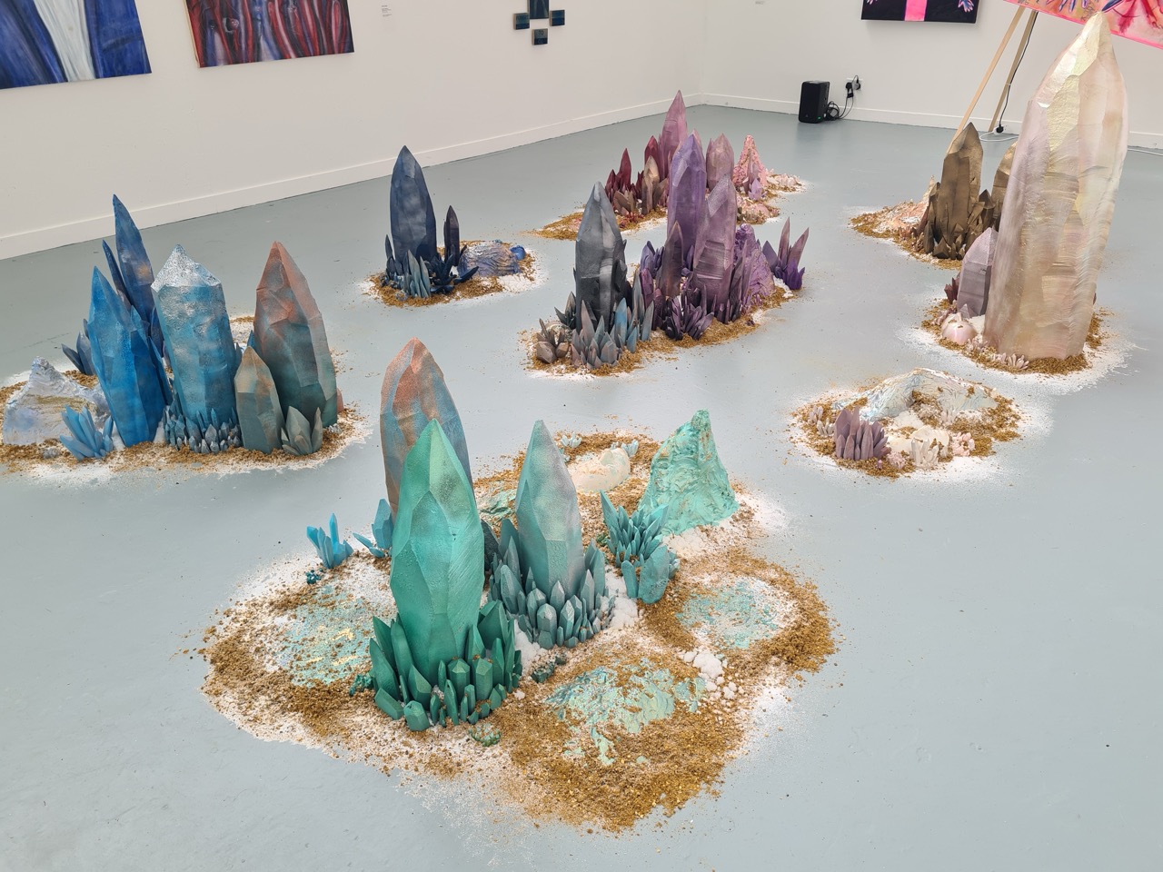 An art installation featuring large, colourful crystal formations of various heights arranged on a light grey floor. Surrounding each crystal are small patches of sand and natural elements, enhancing the artistic display in a gallery setting.