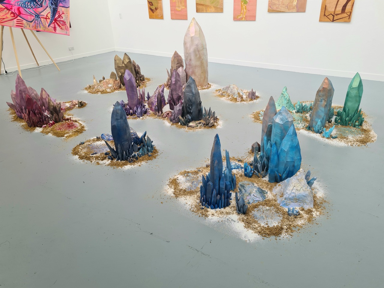 An art installation featuring several large, colourful crystal formations arranged on a grey floor. The crystals vary in colour from purple to blue and are surrounded by small patches of sand, creating a naturalistic landscape in a gallery setting.
