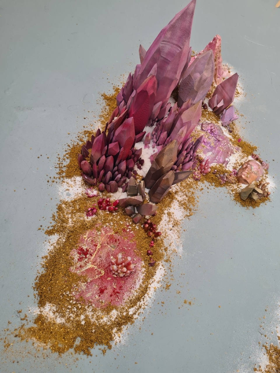 A decorative model depicting a fantasy landscape featuring tall purple crystal formations and clusters of smaller crystals, surrounded by a base of sand and glitter in various shades of pink and gold, set against a light blue surface.