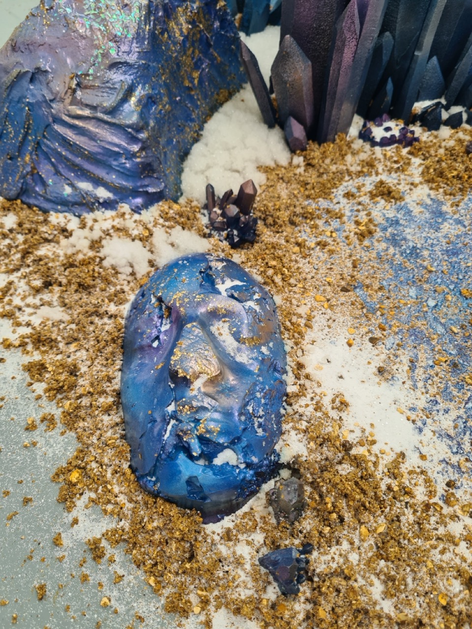 A detailed artistic arrangement featuring a blue and purple face sculpture surrounded by golden glitter and white synthetic crystals. In the background, various crystal structures rise, adding texture to the scene.