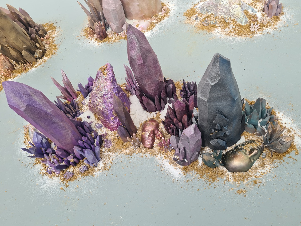 A collection of colourful crystals and gemstones arranged on a light surface. The display includes large purple and grey crystals, smaller clusters in various hues, and scattered golden sand to enhance the overall presentation.