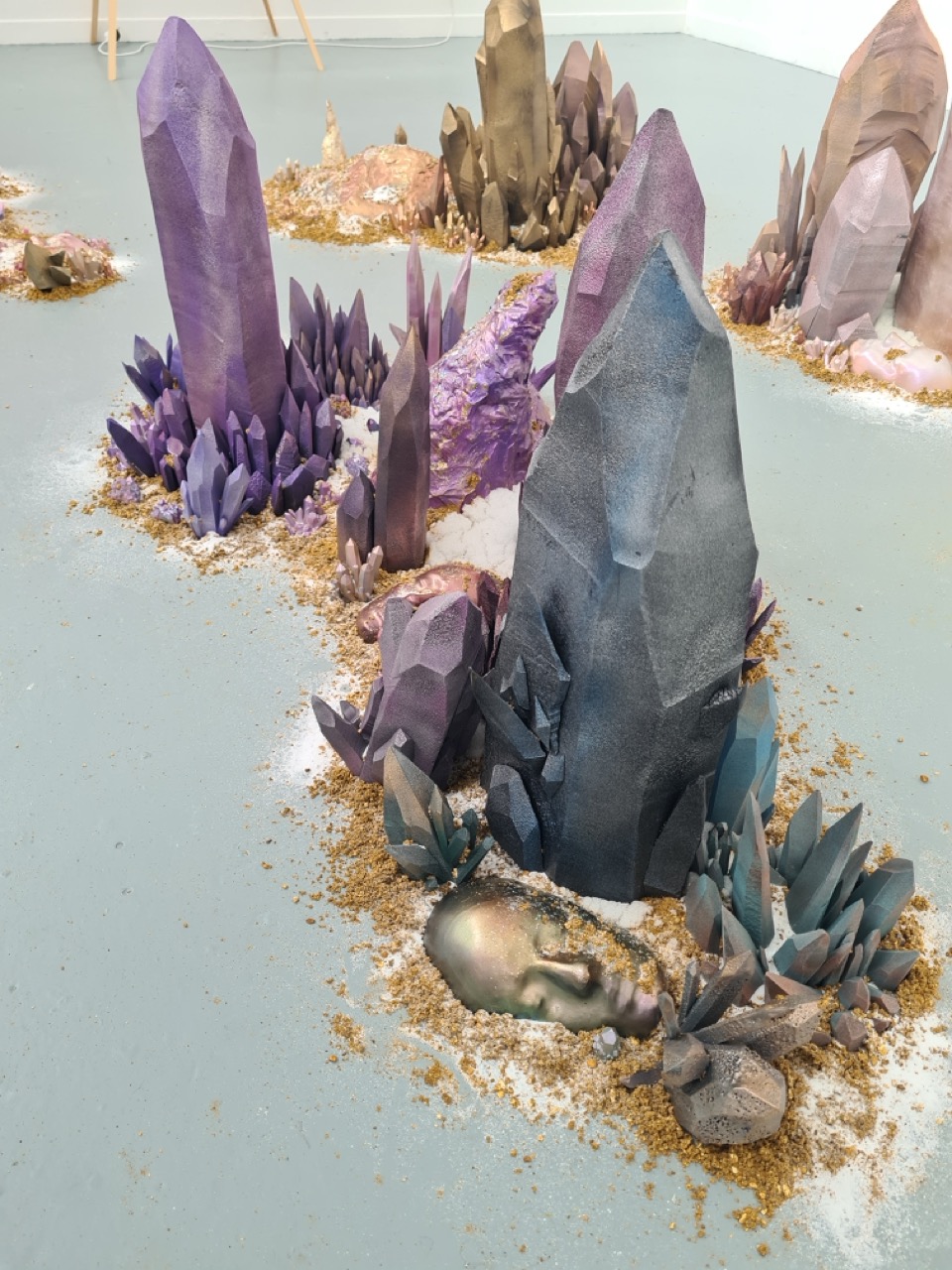 An artistic installation featuring various crystal-like structures in shades of purple, blue, and grey. The crystals are surrounded by earthy materials on a light blue floor, with some sculptures resembling plant forms interspersed throughout the arrangement.