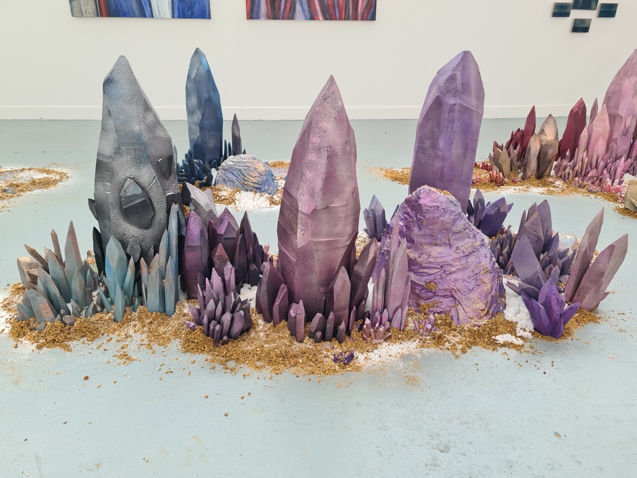 A large installation of artificial crystals in various shades of purple, blue, and grey, arranged on a light-coloured floor. The crystals vary in height and shape, surrounded by scattered golden sand, creating a vibrant display.