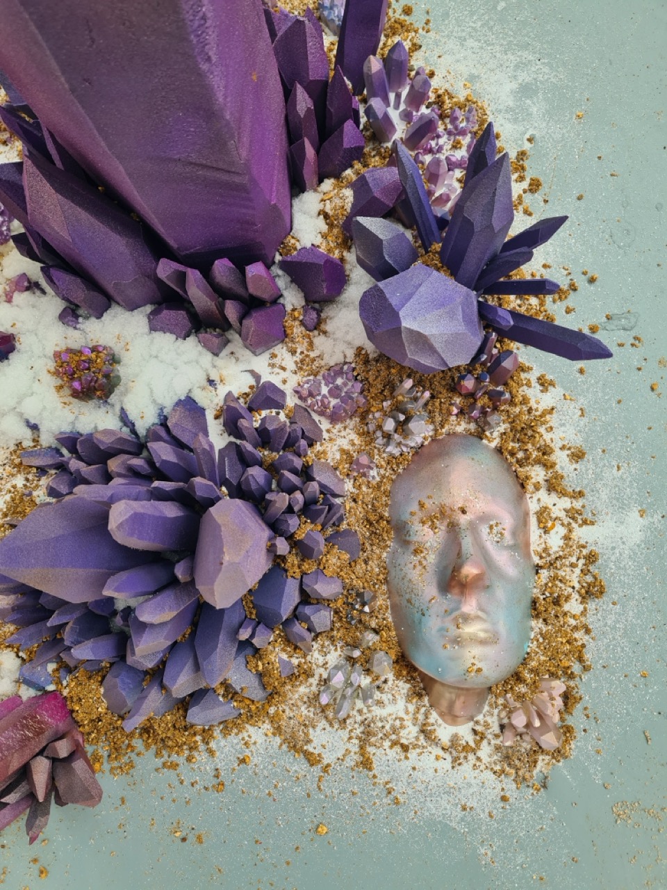 An arrangement of purple crystalline structures and flowers surrounded by golden sand, with a metallic face mask resting in the centre. The background is a light blue surface that enhances the vibrant colours of the crystals and the mask.