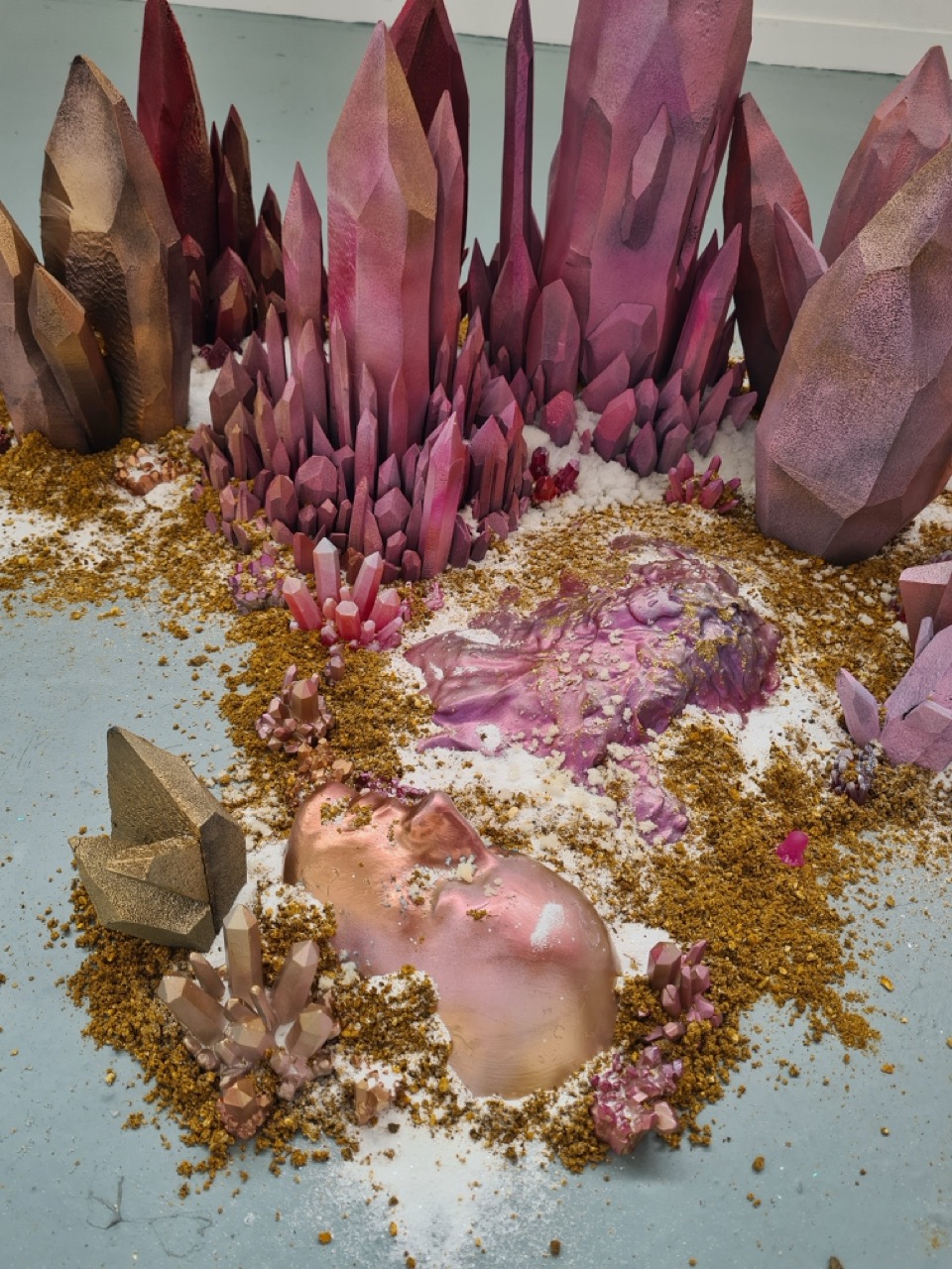 An artistic arrangement featuring large pink and purple crystalline structures accompanied by smaller crystals and golden sand. The crystals vary in height and shape, with a smooth, human-like form partly buried in the sand, surrounded by additional small crystal formations.