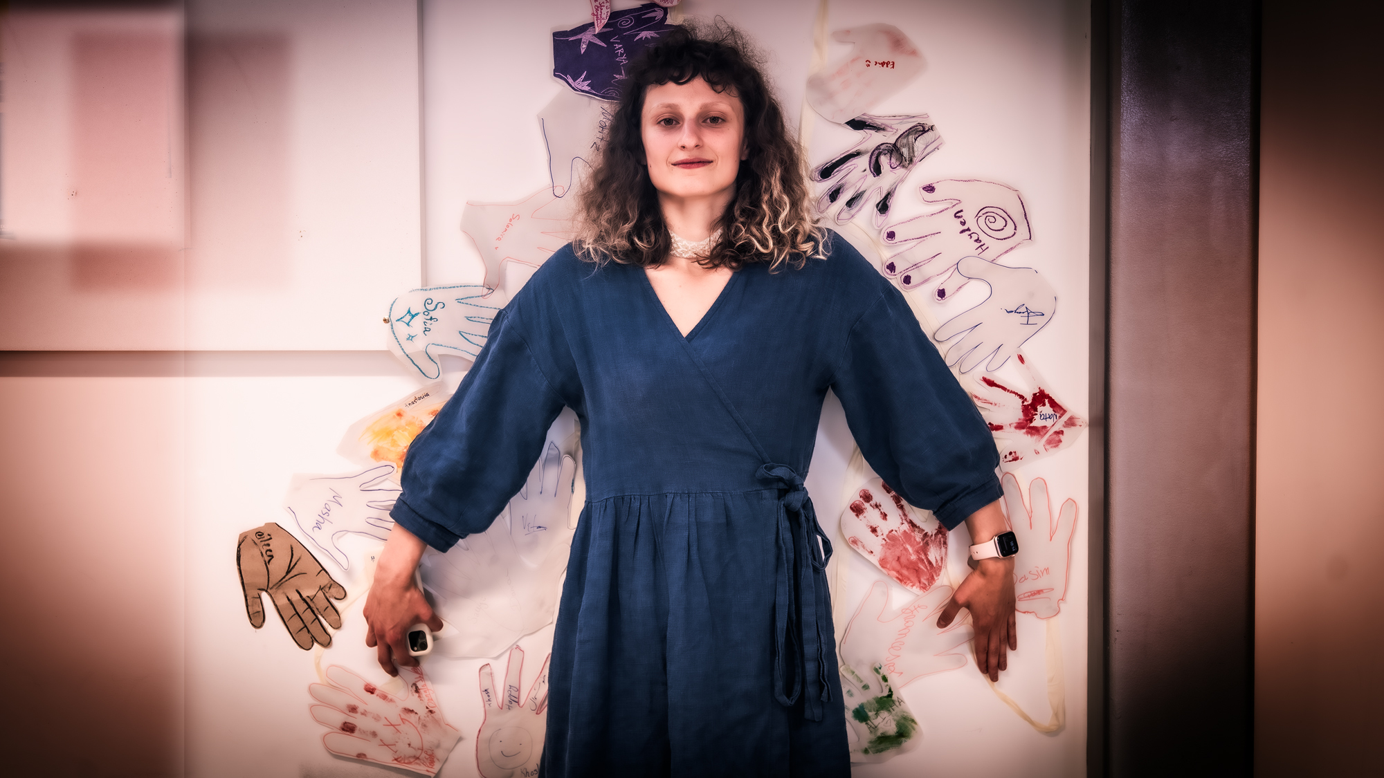 Woman in navy blue dress with purple hair accessory standing against white wall with sketched drawings and coloured elements.