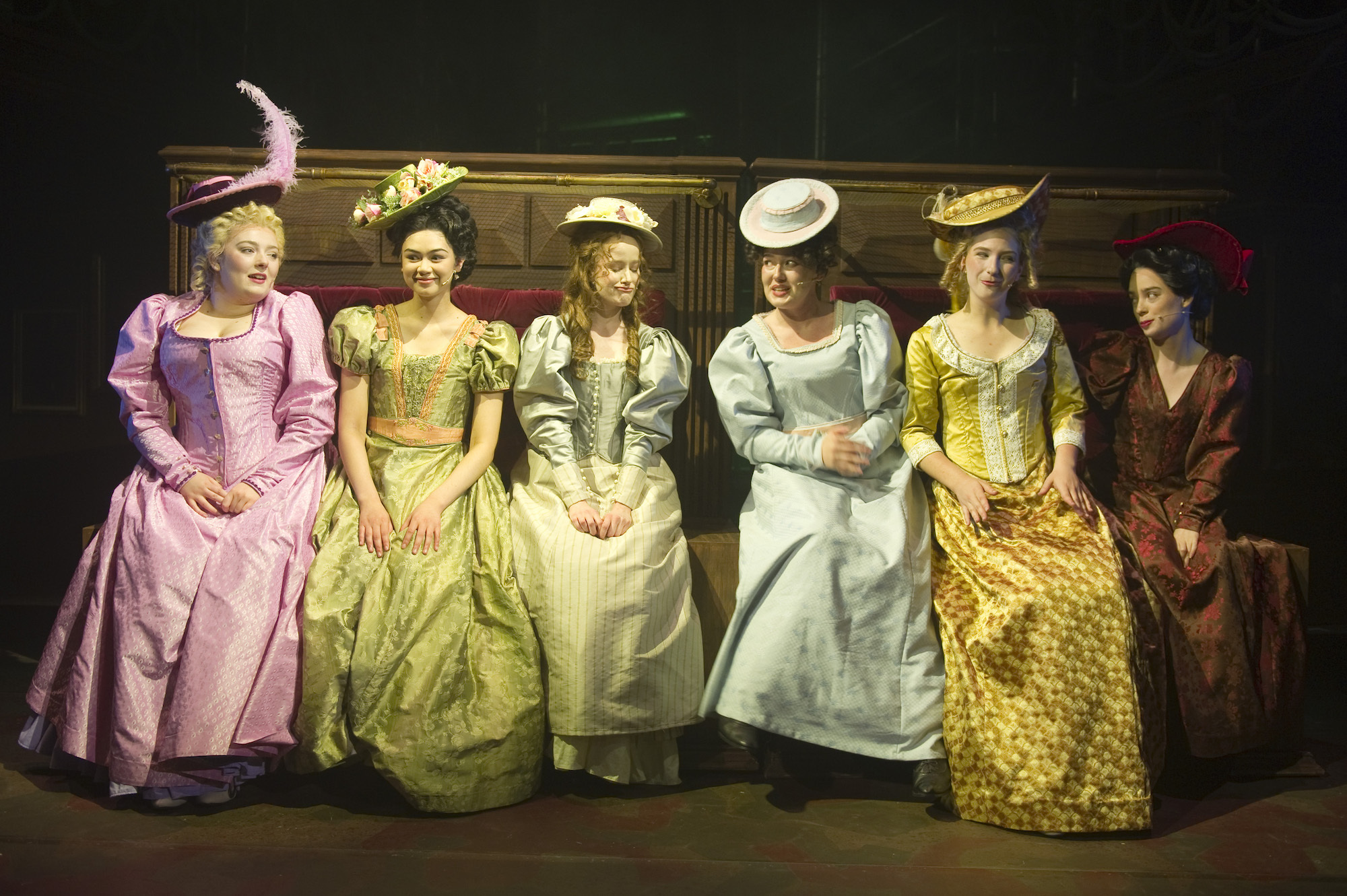 Six BA (Hons) Acting students sitting along a high-backed wooden bench, all wearing period-style costumes while performing AUB Productions' "Masque Macabre".