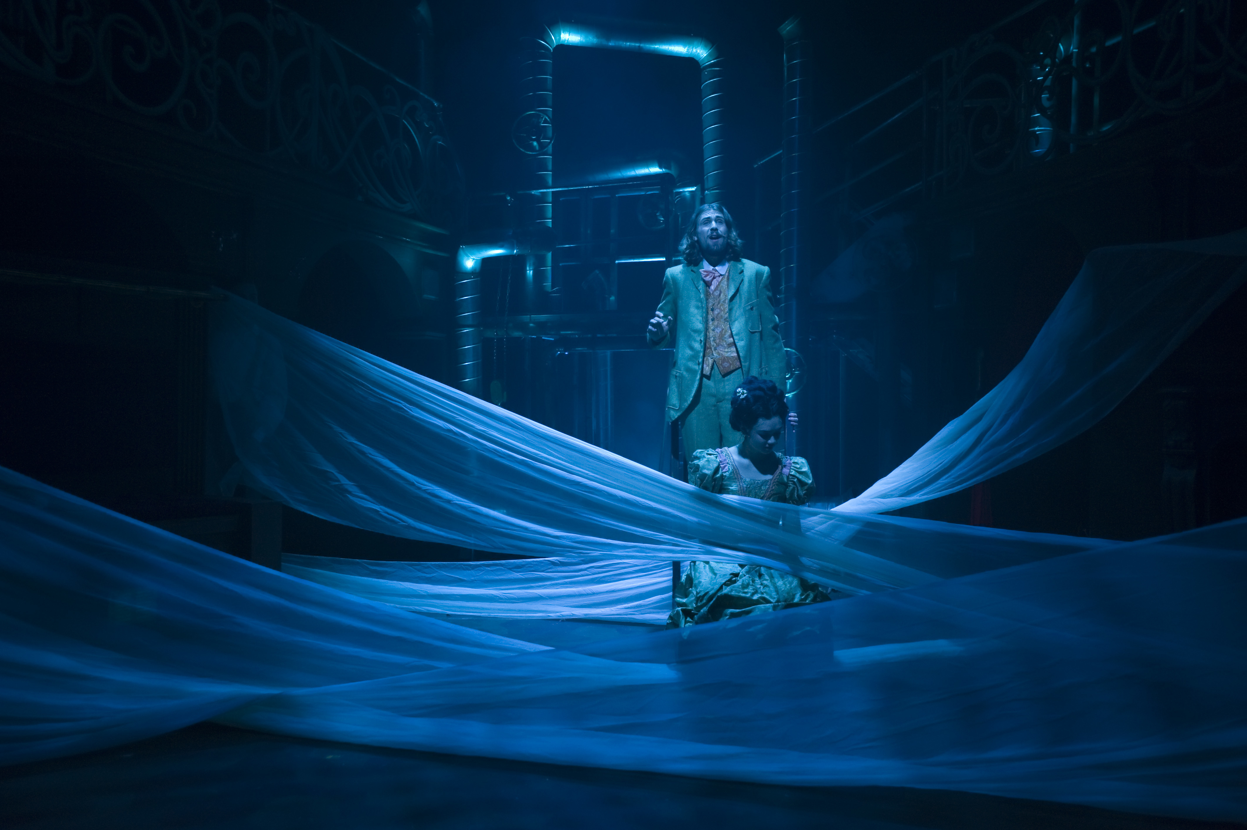 Two BA (Hons) Acting students on a dimly lit stage in the middle of a performance of "Masque Macabre". One actor is standing, the other is sitting in front of him. Lengths of material are draped across the stage between them.