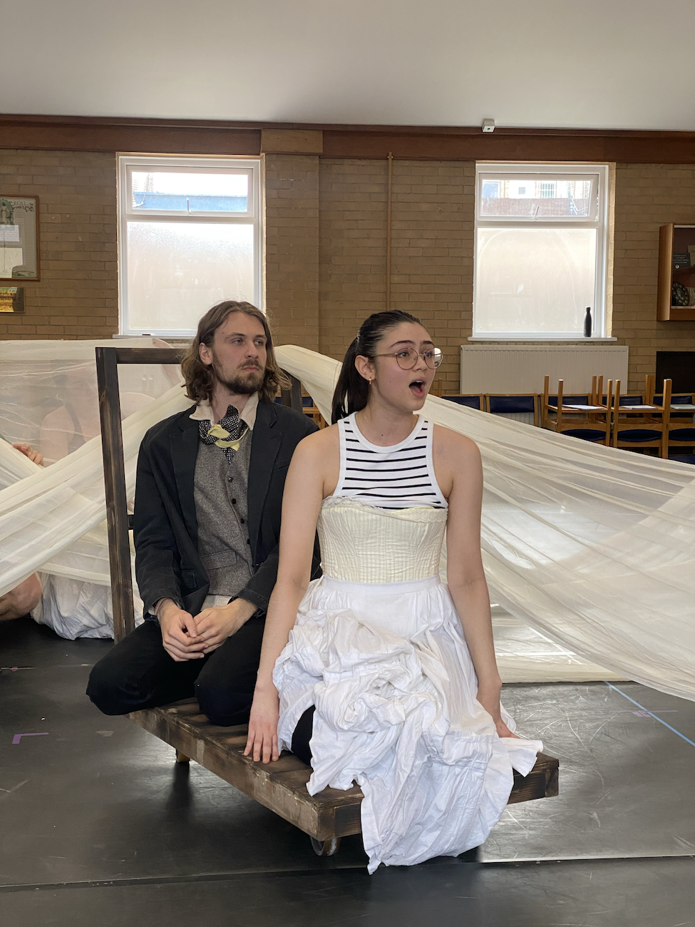 Two BA (Hons) Acting students, one in full costume, the other in partial costume, kneeling on a trolley while in rehearsal for Masque Macabre.