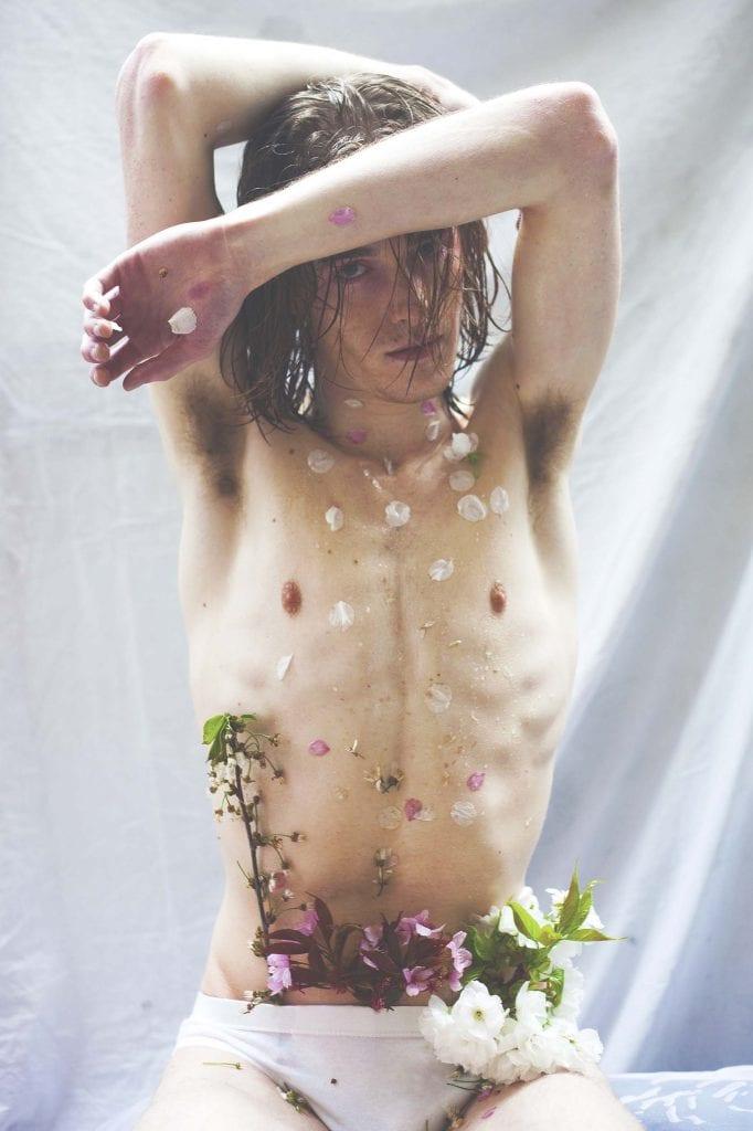 A shirtless model raises their arms above their head with flower petals falling down in front of them.