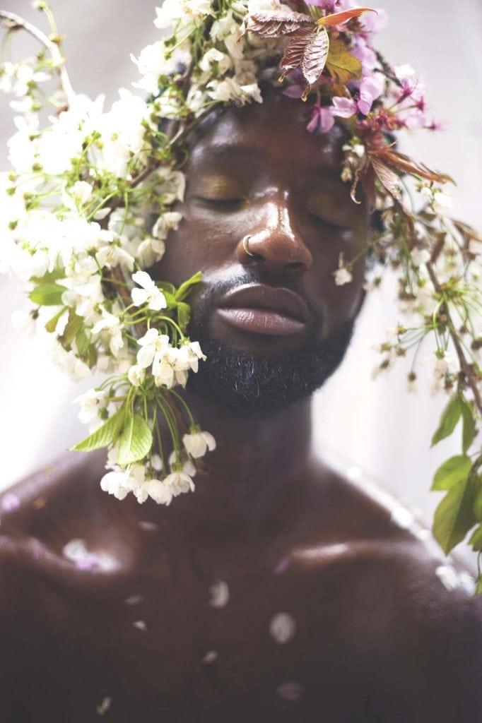 Close up of a shirtless model with their eyes closed and plants and flowers on their head.