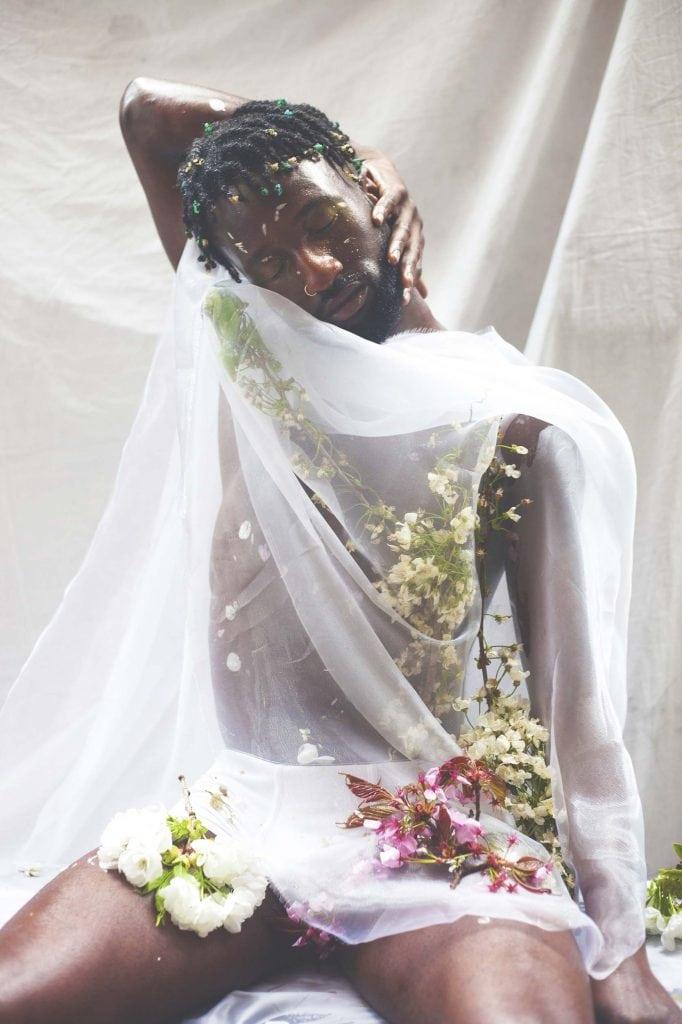 Shirtless model with translucent white material draped over them poses for a photo with their eyes closed.
