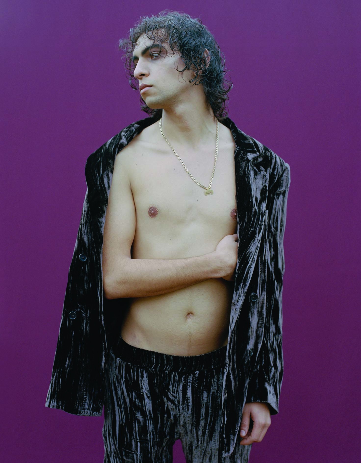 Shirtless model with a black suede jacket held over their shoulders standing against a purple background.